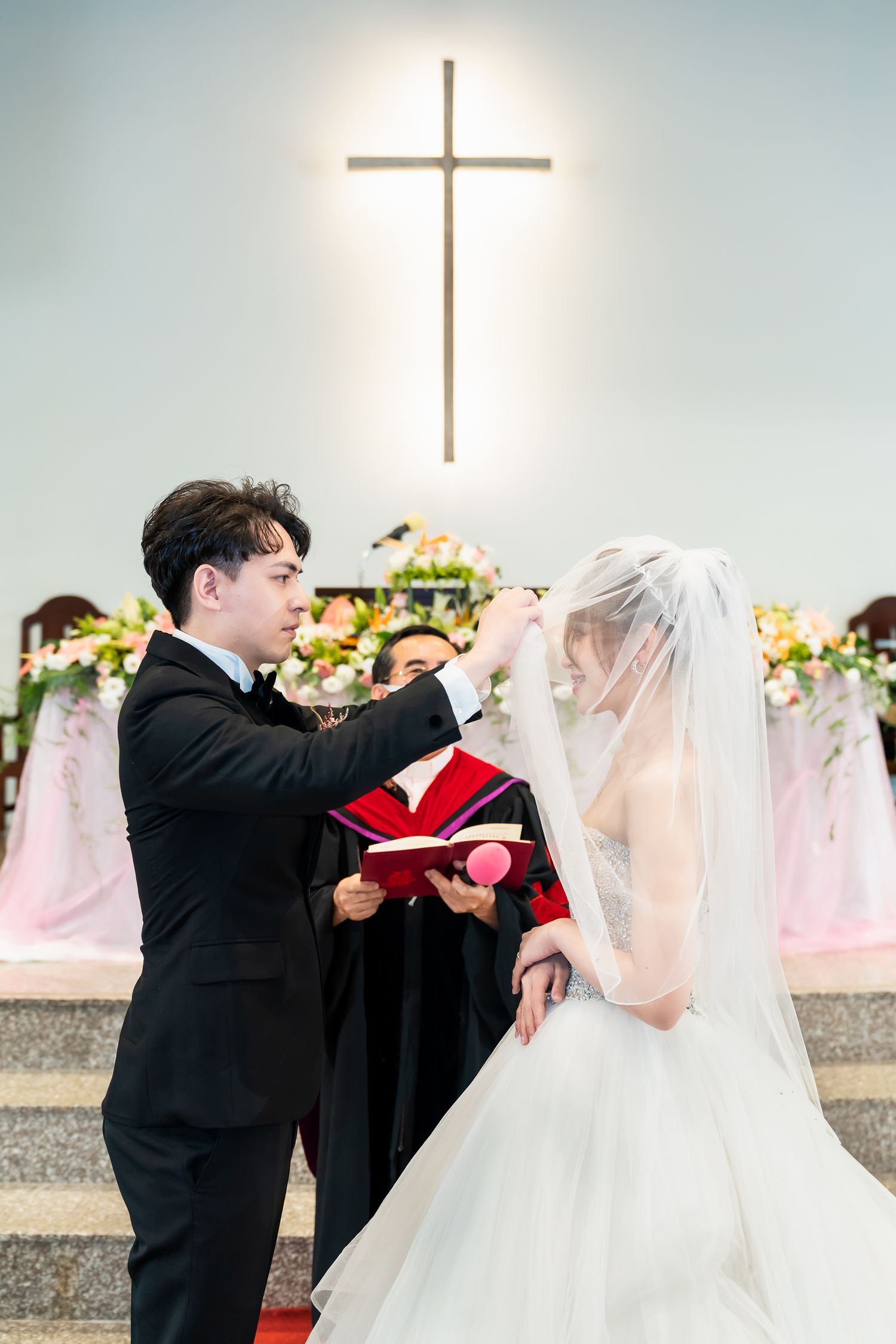 A wedding ceremony with the groom lifting the bride's veil beneath an illuminated cross.