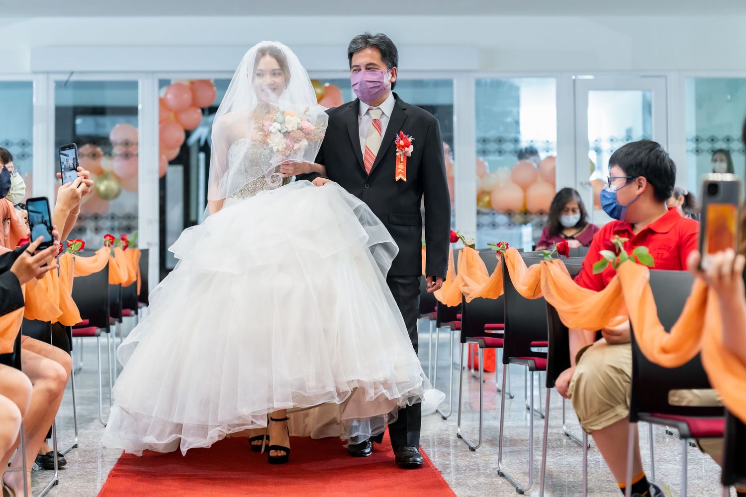 Wedding celebration on red carpet with guests watching in chairs with orange decorations.
