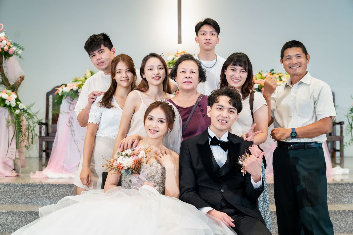 Wedding group photo showing joyful celebration with decorative floral arch in background.