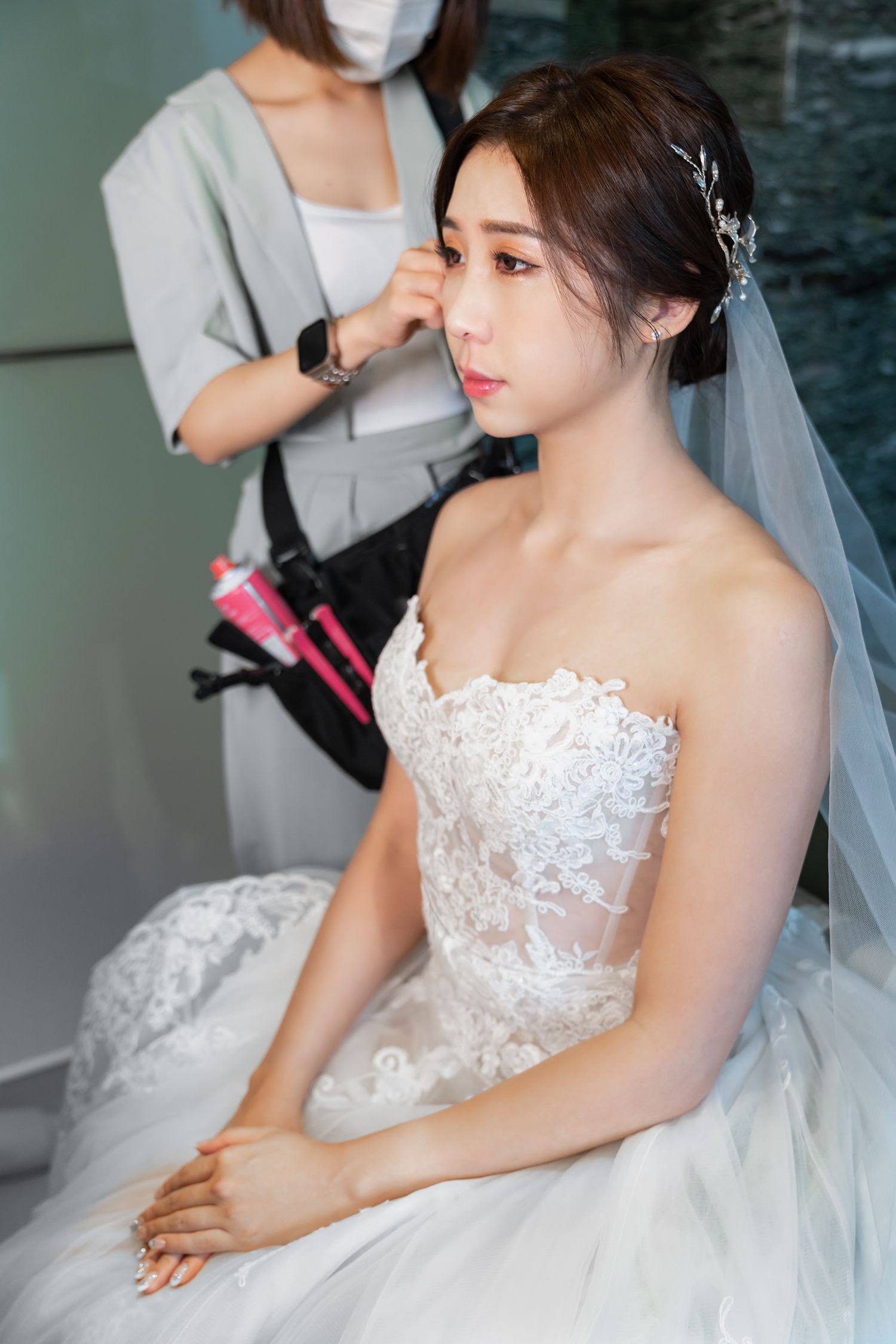 A bride in a white lace wedding dress and veil sits while having her hair styled during wedding preparations.