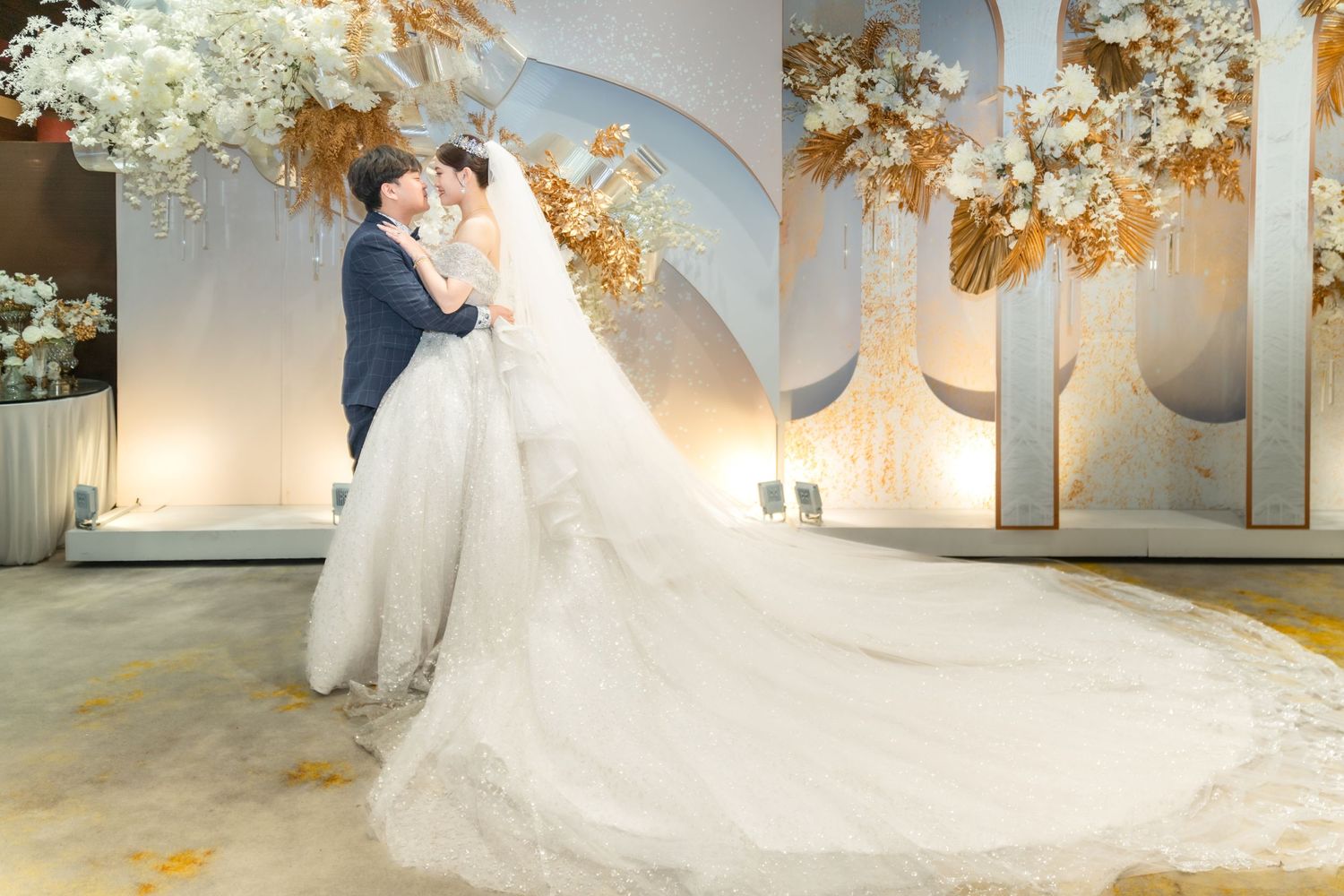 A couple embraces at their wedding ceremony under an elegant arch decorated with white flowers and dried pampas grass.