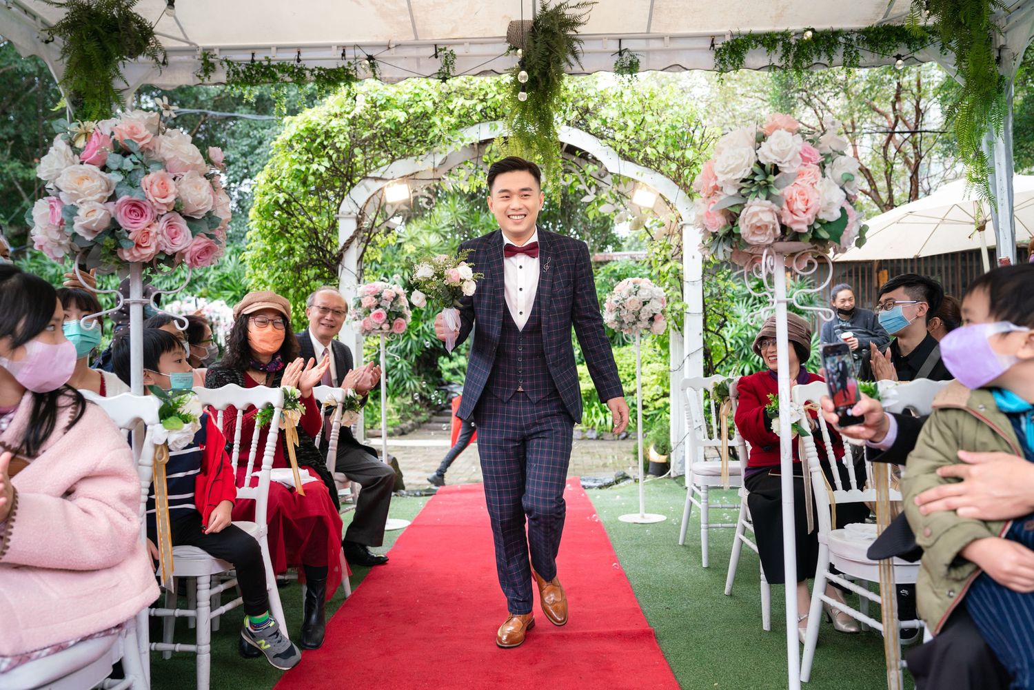 A formal outdoor wedding ceremony features an elegant walkway with a floral arch and red carpet surrounded by seated guests.