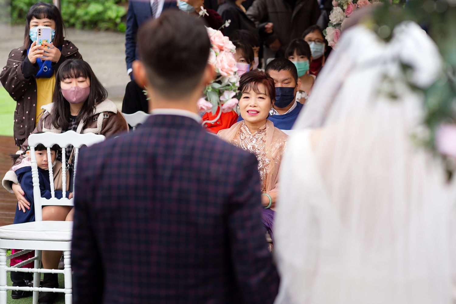 Wedding guests watch a ceremony outdoors while some capture photos on their phones during a sunny day.