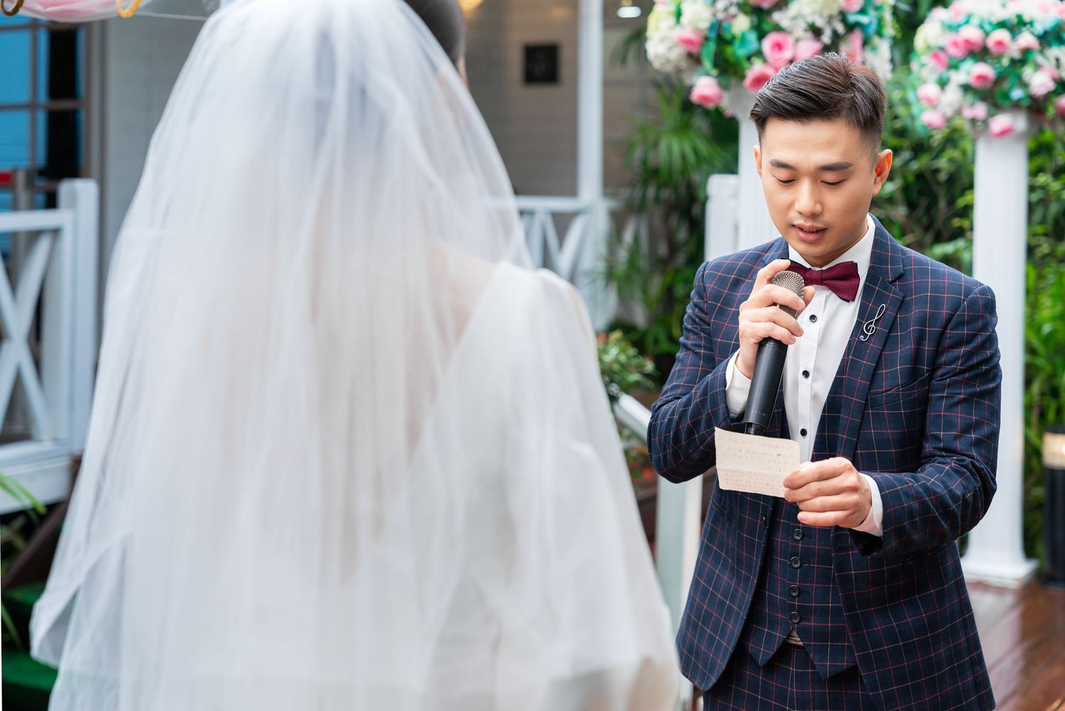Person in plaid suit reading vows from paper during outdoor garden wedding ceremony with bride in white dress and veil.