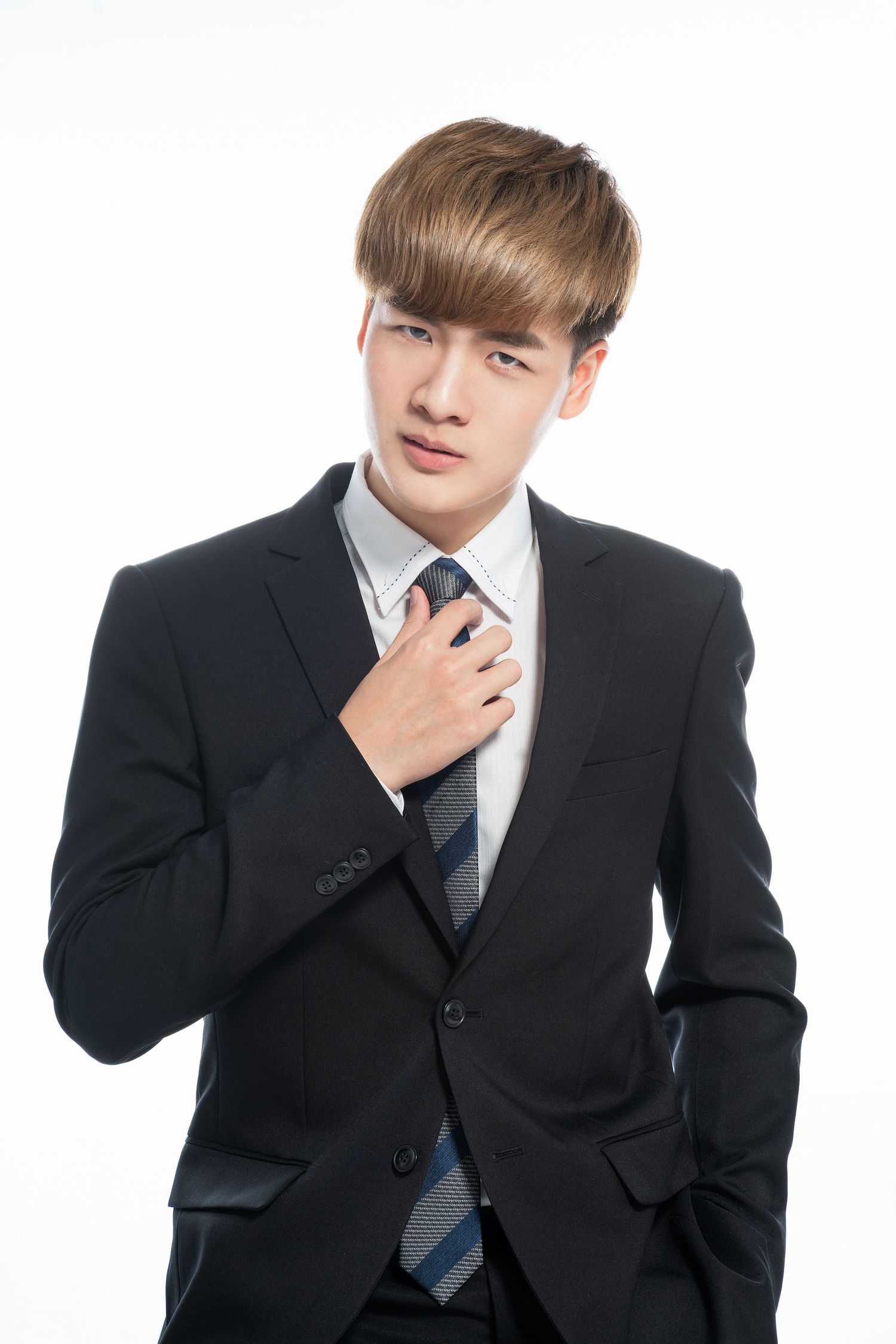 Young professional in black suit and striped tie adjusts collar while posing against white background.