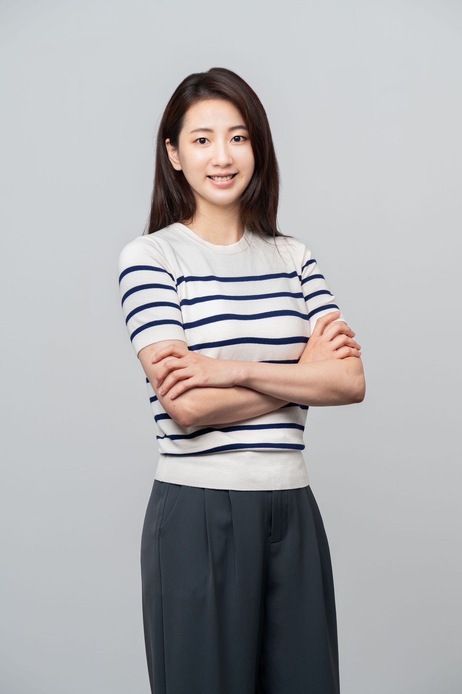 Person in striped crewneck sweater and dark trousers standing with arms crossed against gray background.