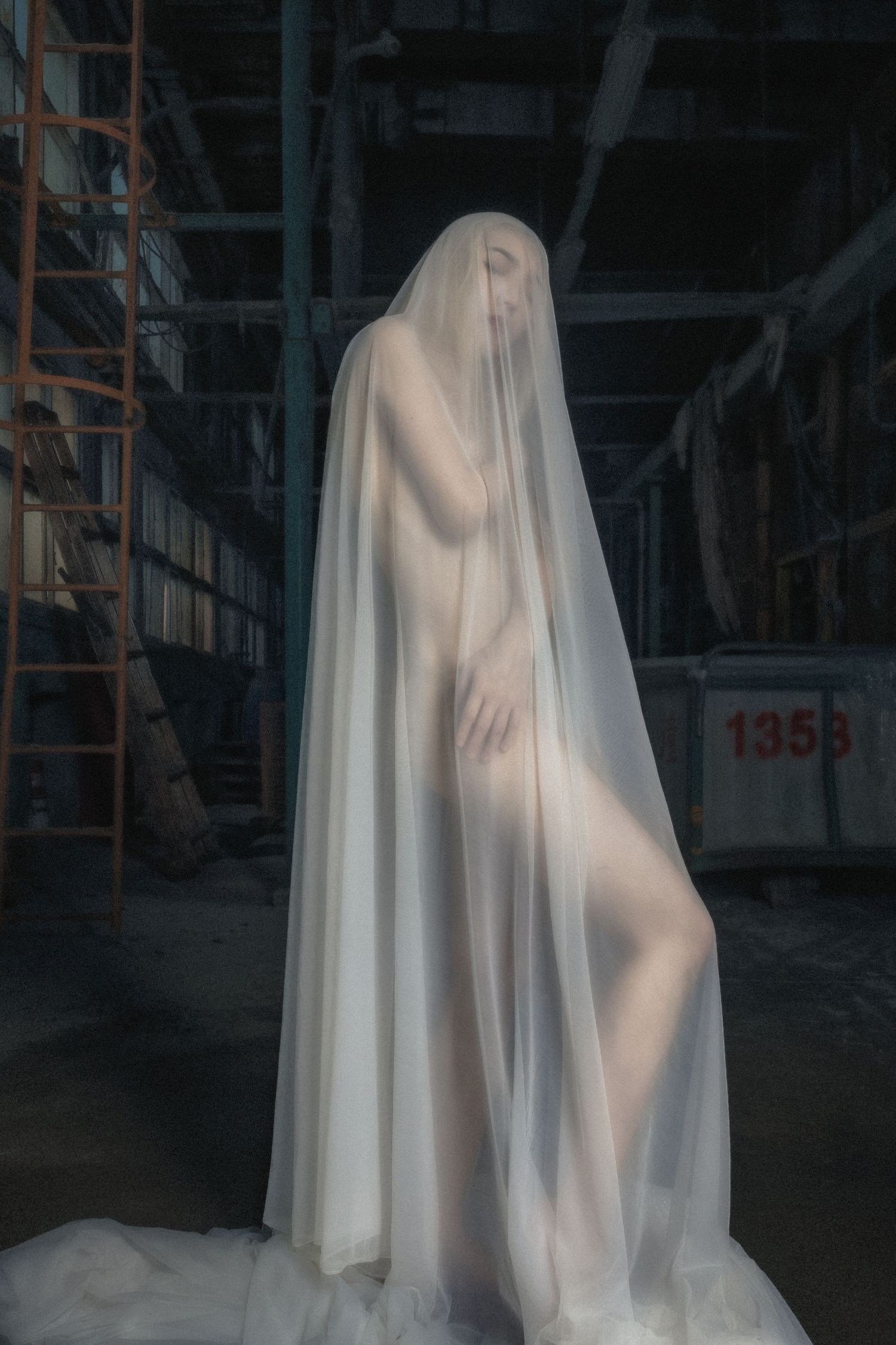 A figure draped in ethereal white fabric stands in an abandoned industrial space creating a ghostly silhouette.