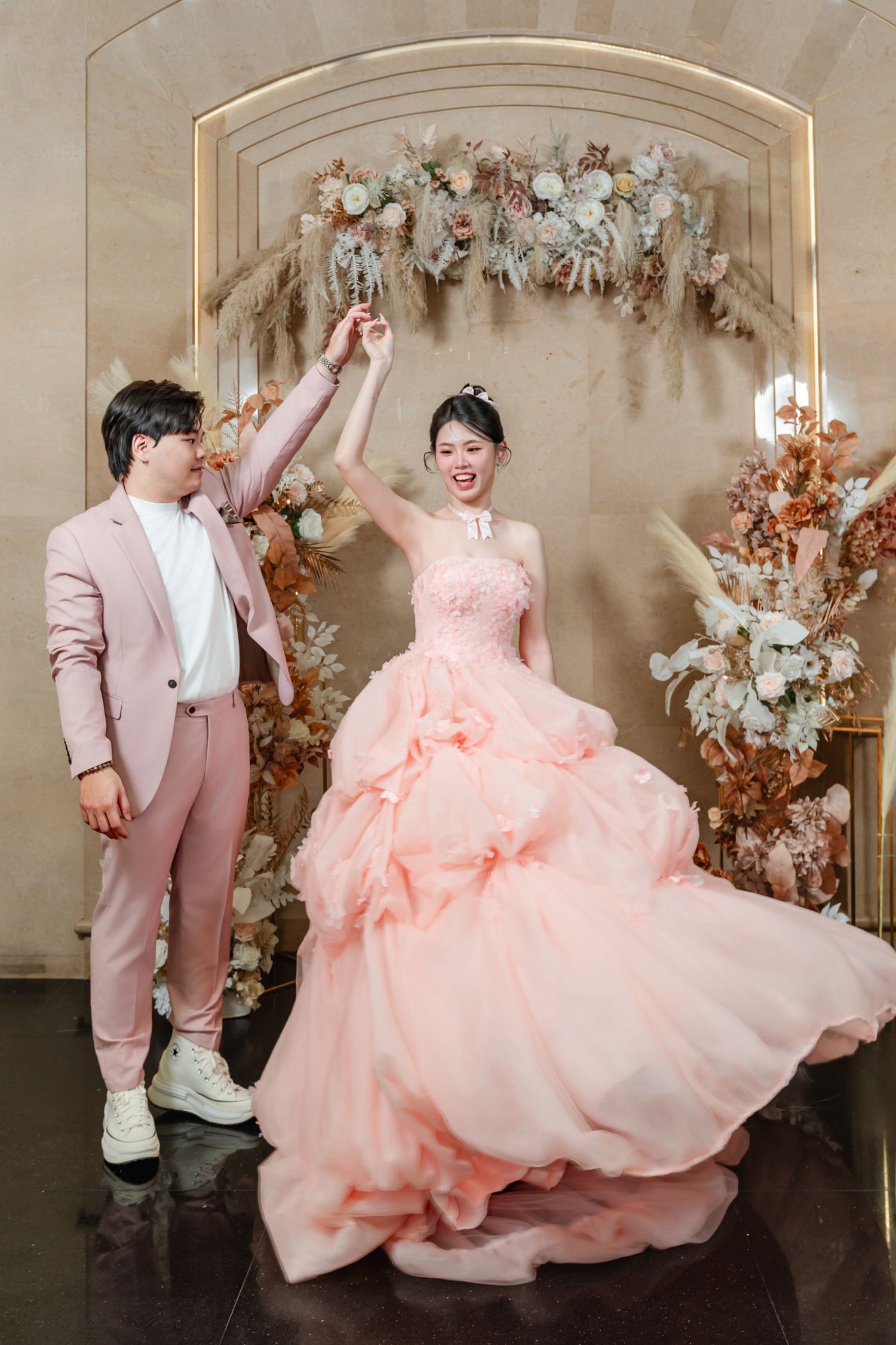 A joyful couple in matching pink attire pose under a floral arch at their wedding celebration.
