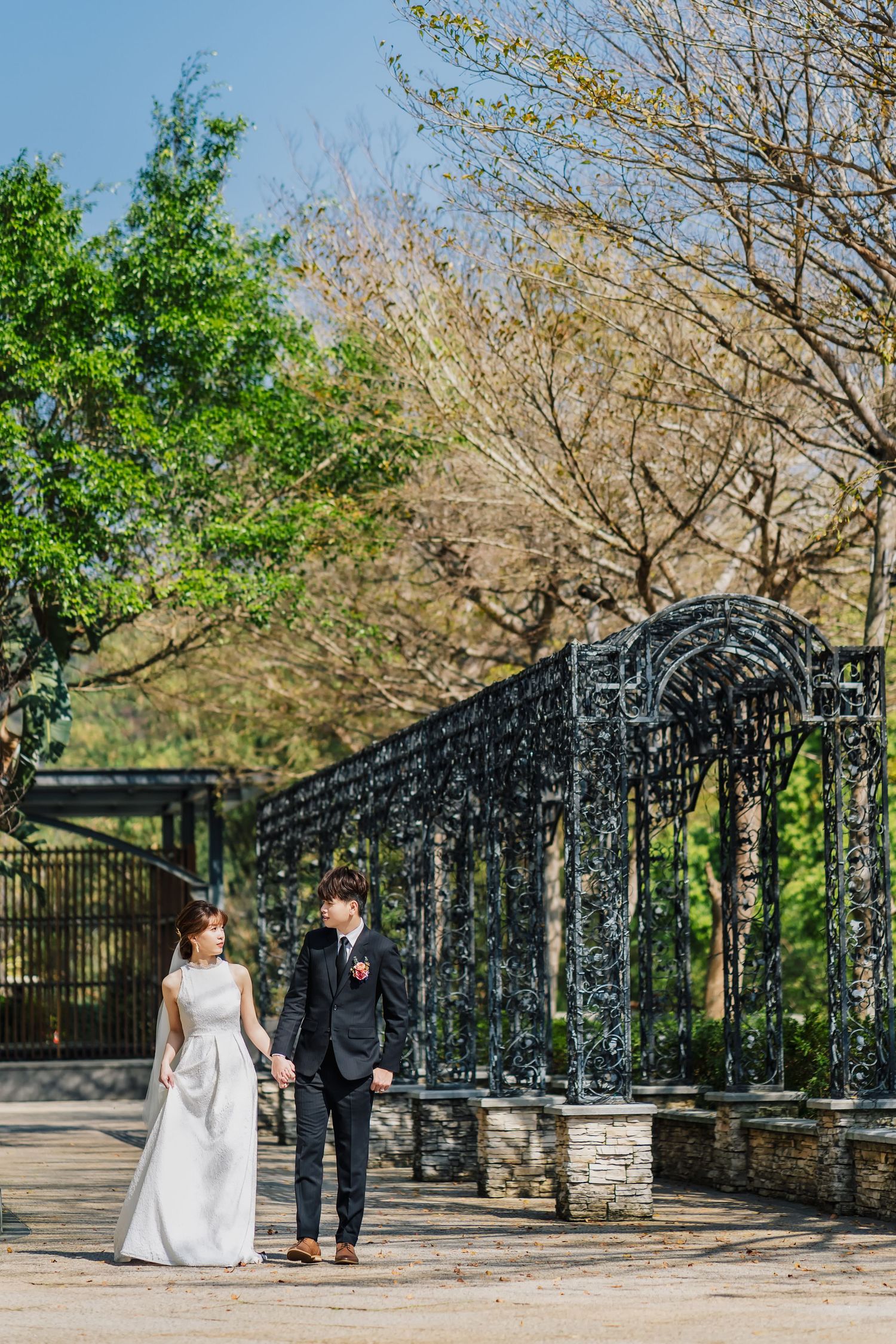 A wedding couple stands together on a path by an ornate black iron archway and stone wall on a sunny spring day.