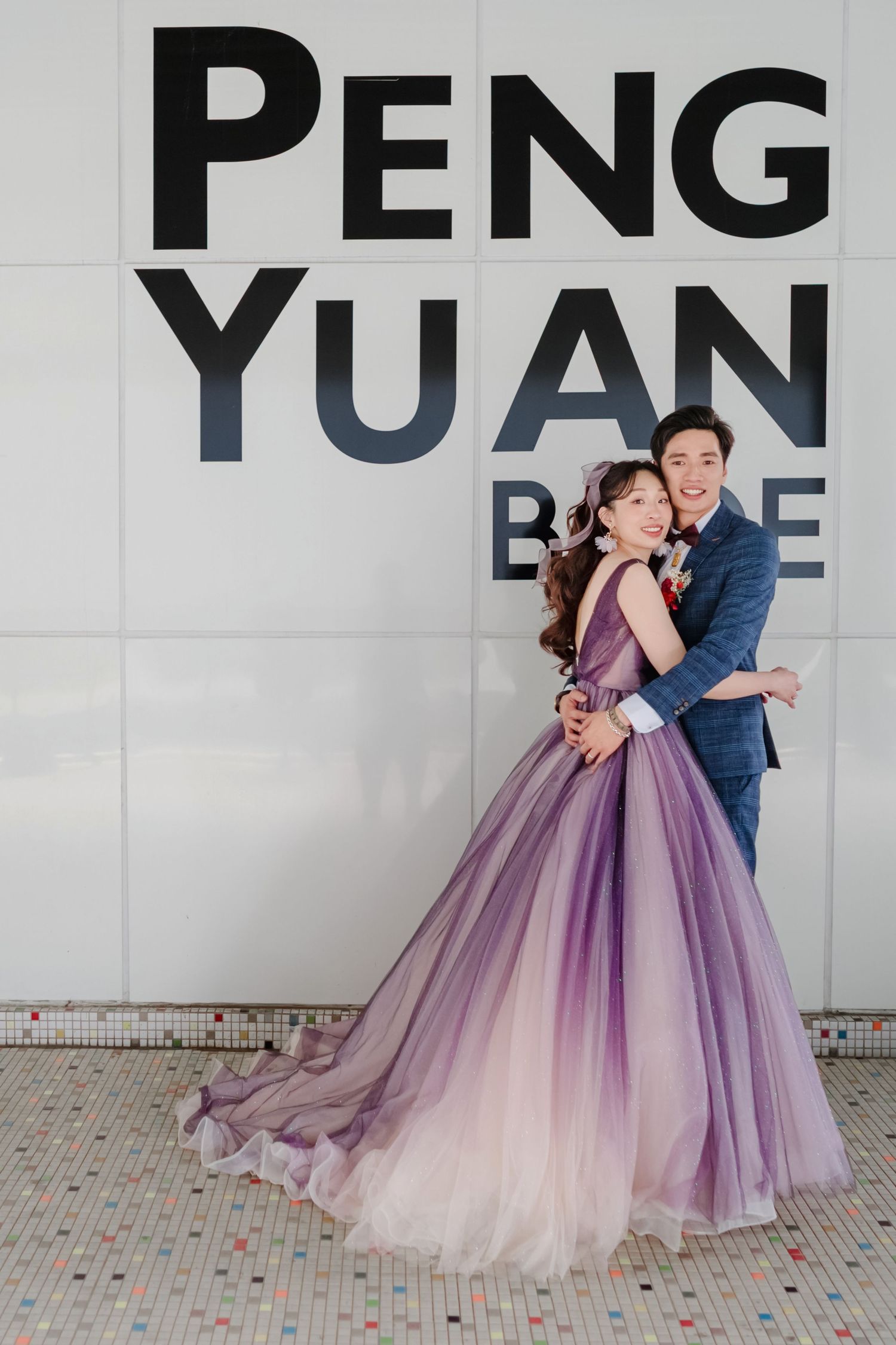 Elegant couple in formal attire embrace in front of PENG YUAN sign, with bride wearing flowing purple ombre gown.