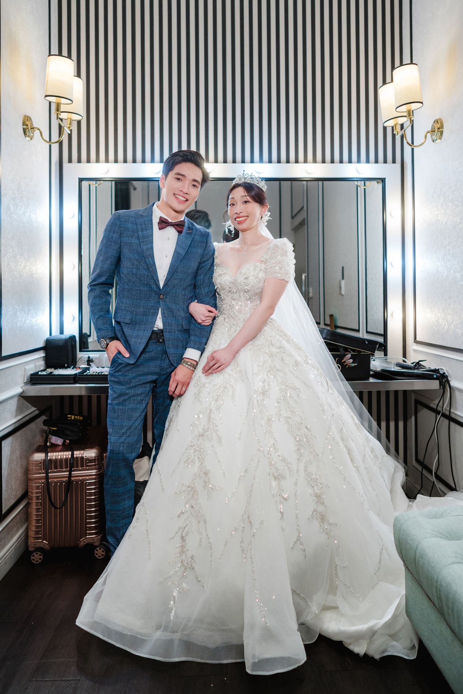 A couple poses together in wedding attire - a blue suit and bow tie paired with an elegant white lace wedding gown.