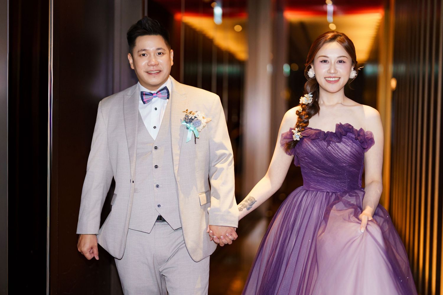 A couple in formal attire walking together, with the groom in a light gray suit and bride in a purple evening gown.