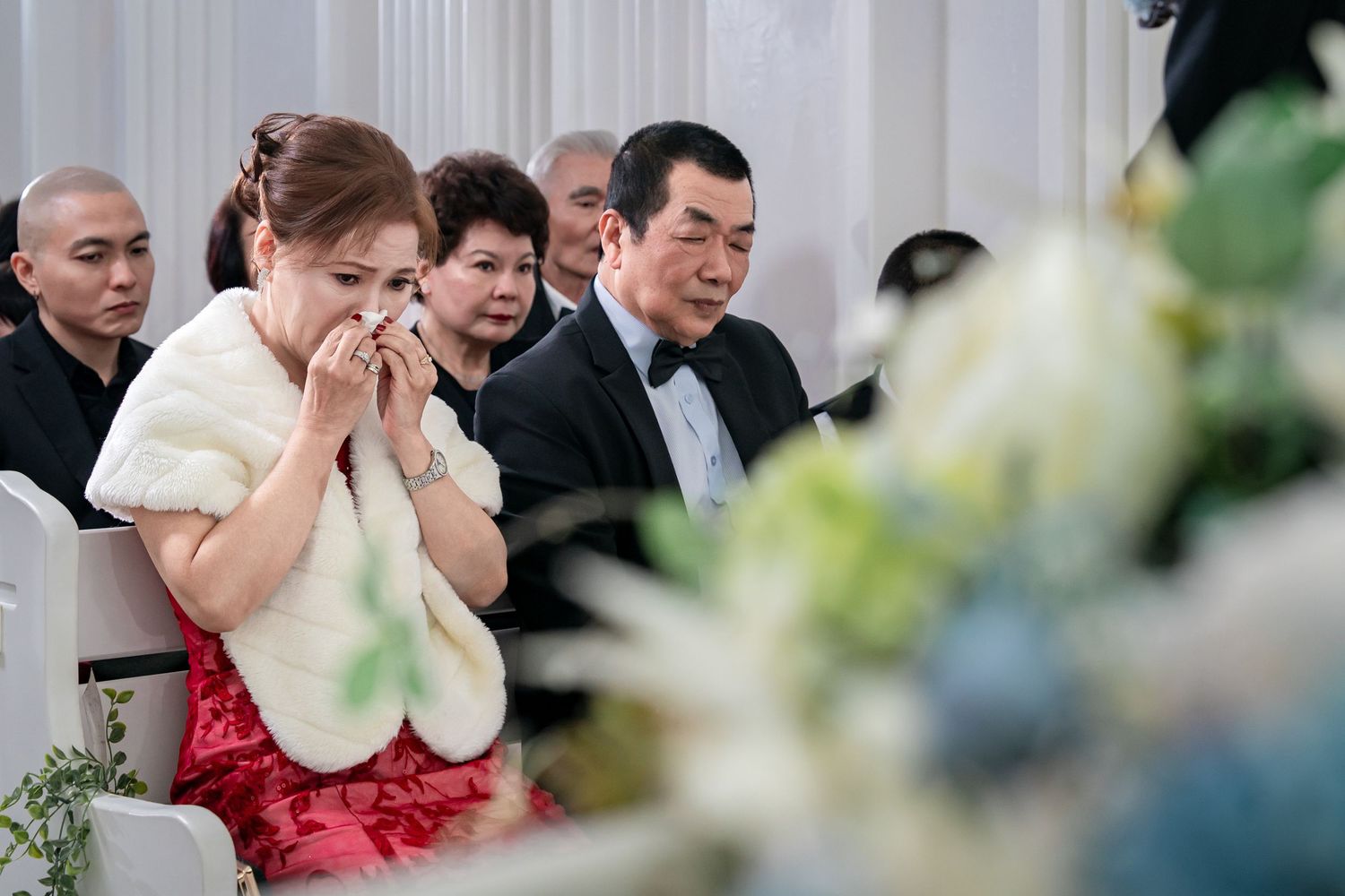 Guests at a wedding ceremony show heartfelt emotions in a bright white venue with chandeliers.
