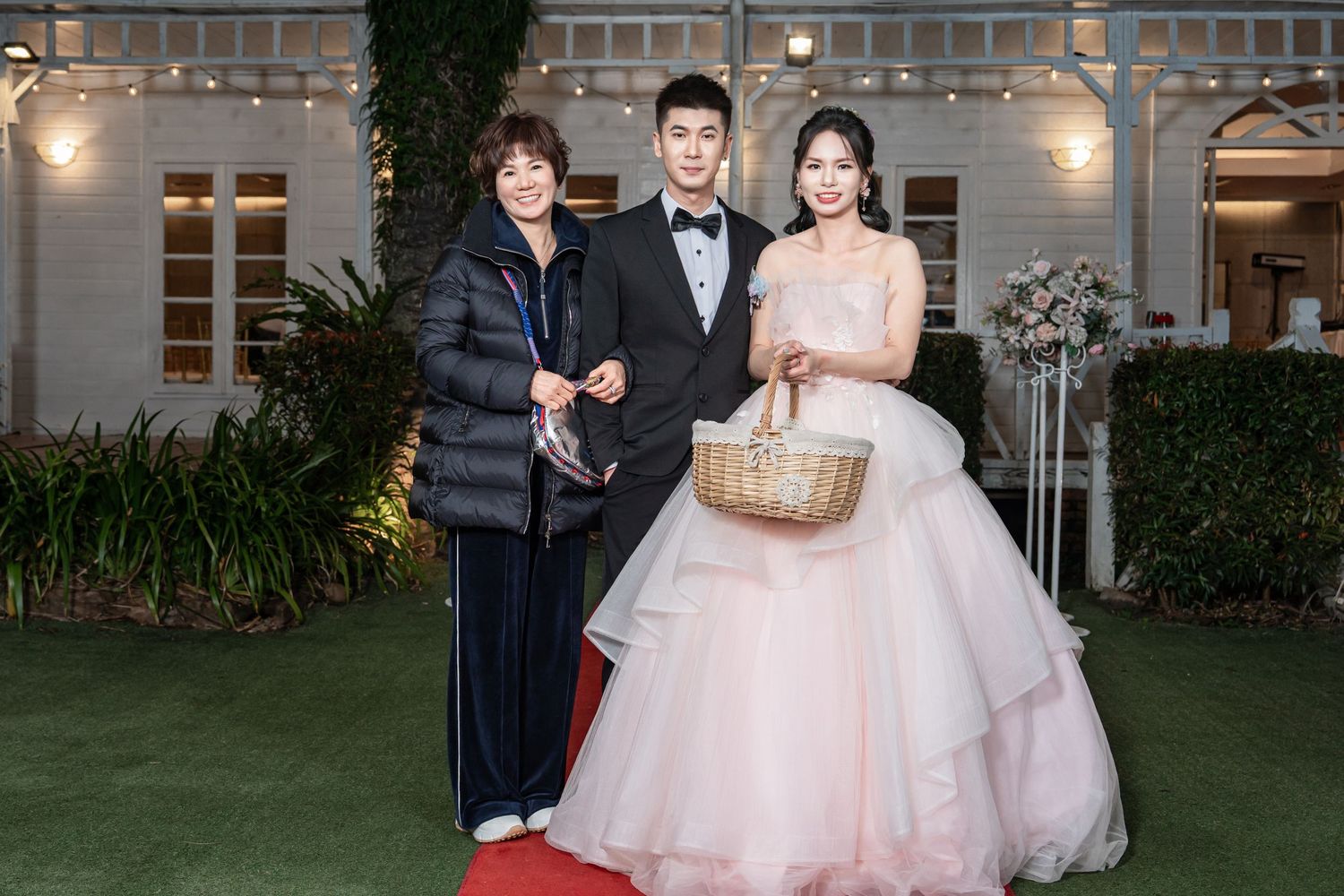 A group in formal attire poses together at an outdoor wedding venue with decorative lighting and greenery in the background.