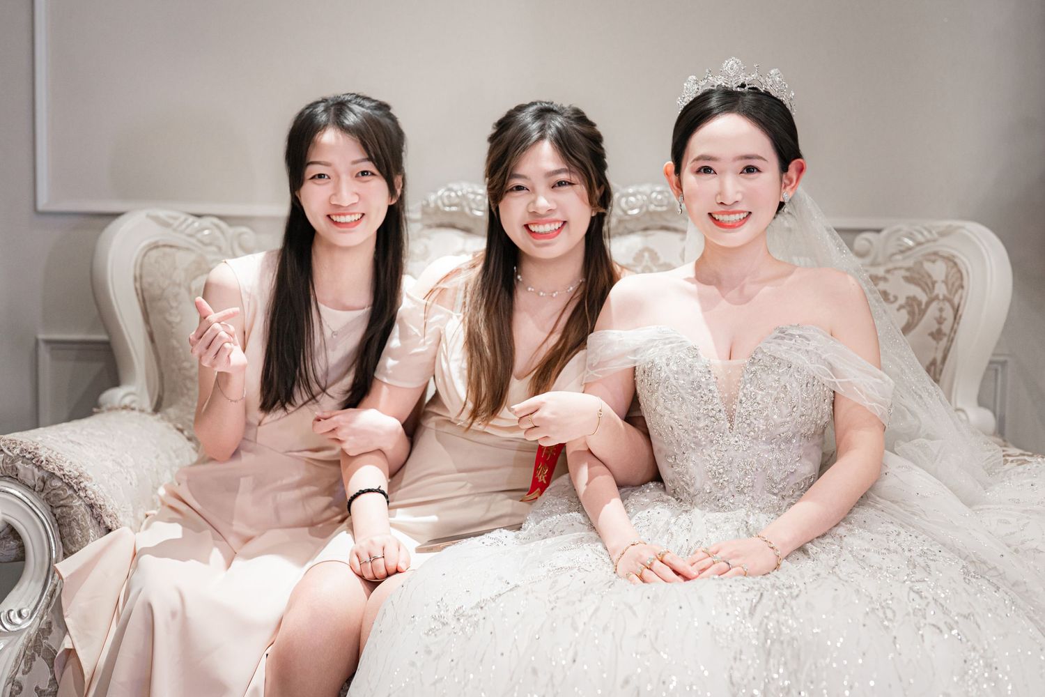 Three women in elegant pink and white formal dresses sit together smiling on an ornate white sofa.