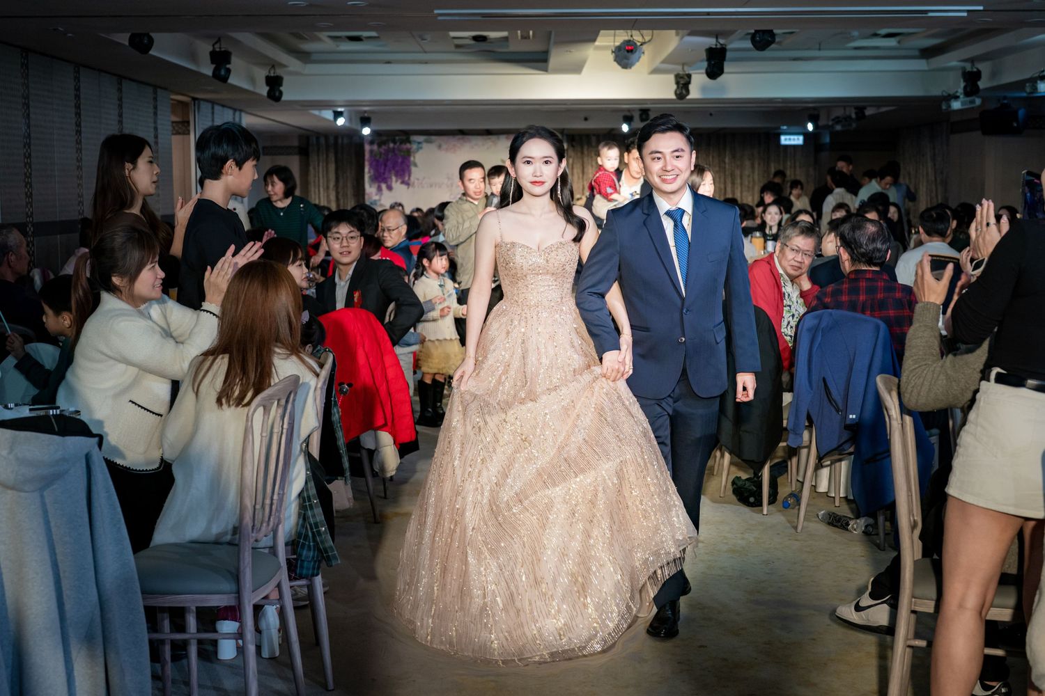 Couple walking down the aisle at a wedding reception in a modern venue with guests looking on from both sides.