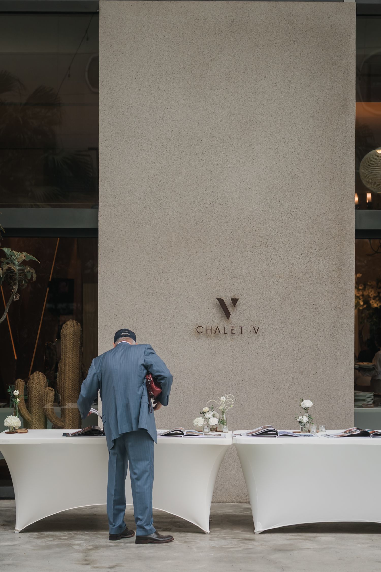 A blue-uniformed worker sets up dining tables against a white wall with 'CHALET V' text in an elegant restaurant setting.
