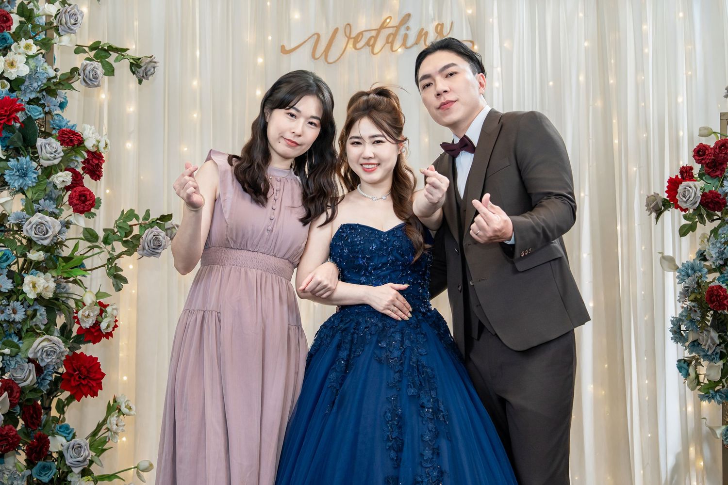 Three people pose at a wedding celebration backdrop with red roses and white curtains.