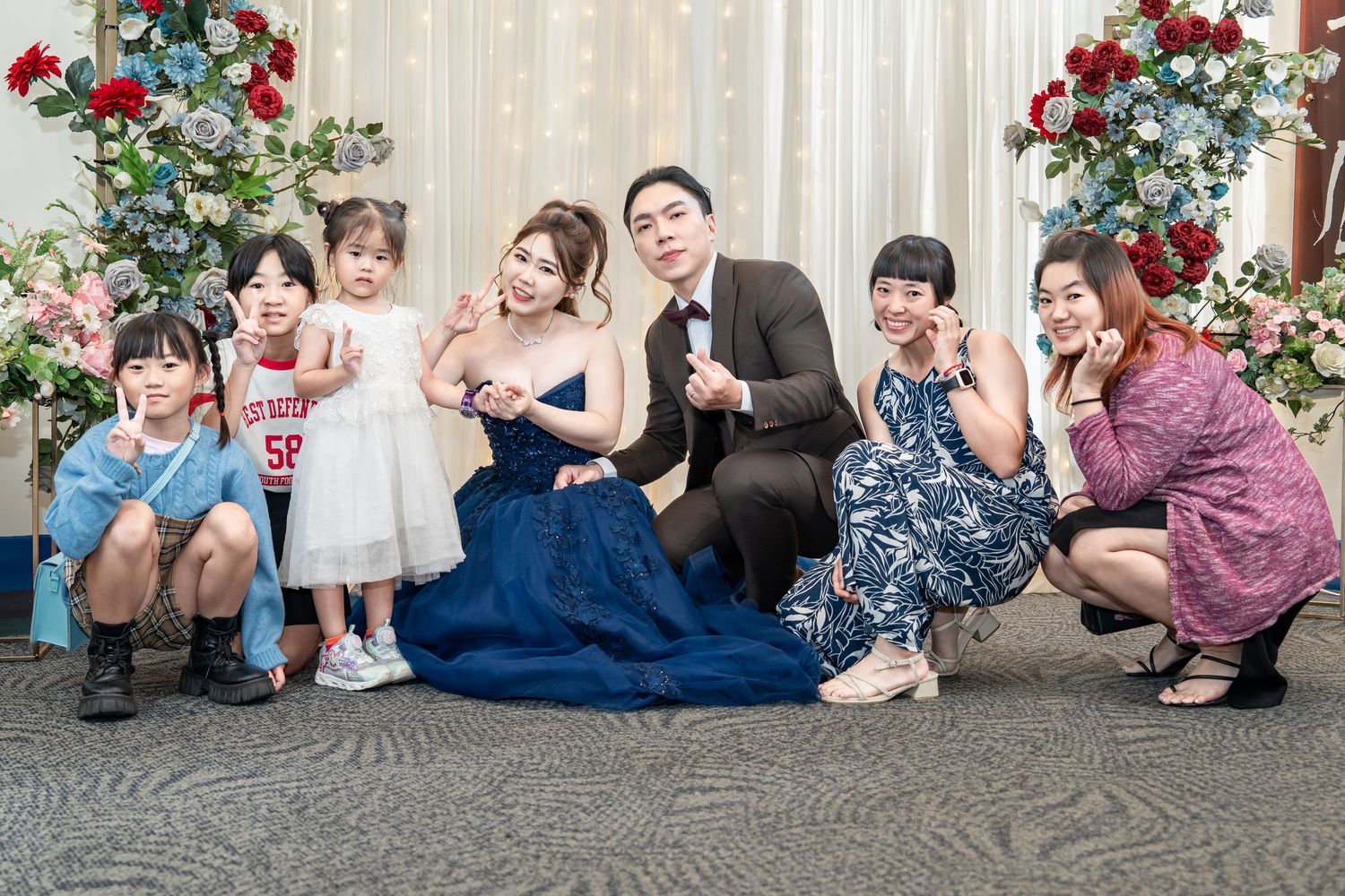A group of people pose together against a beige wall with red roses and greenery decorations.