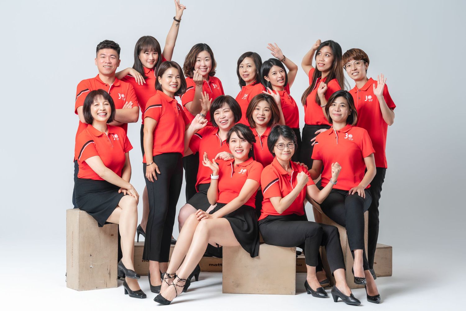 A group in matching red polo shirts and black pants pose together on wooden boxes in a studio setting.