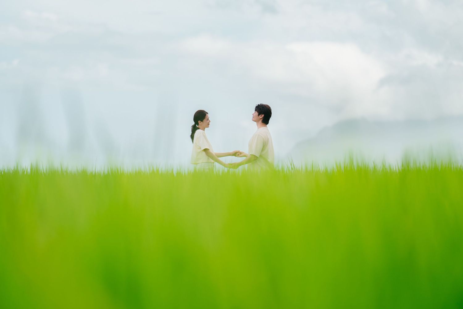 Two people sit together in tall green grass on a warm summer day with a cloudy sky overhead.
