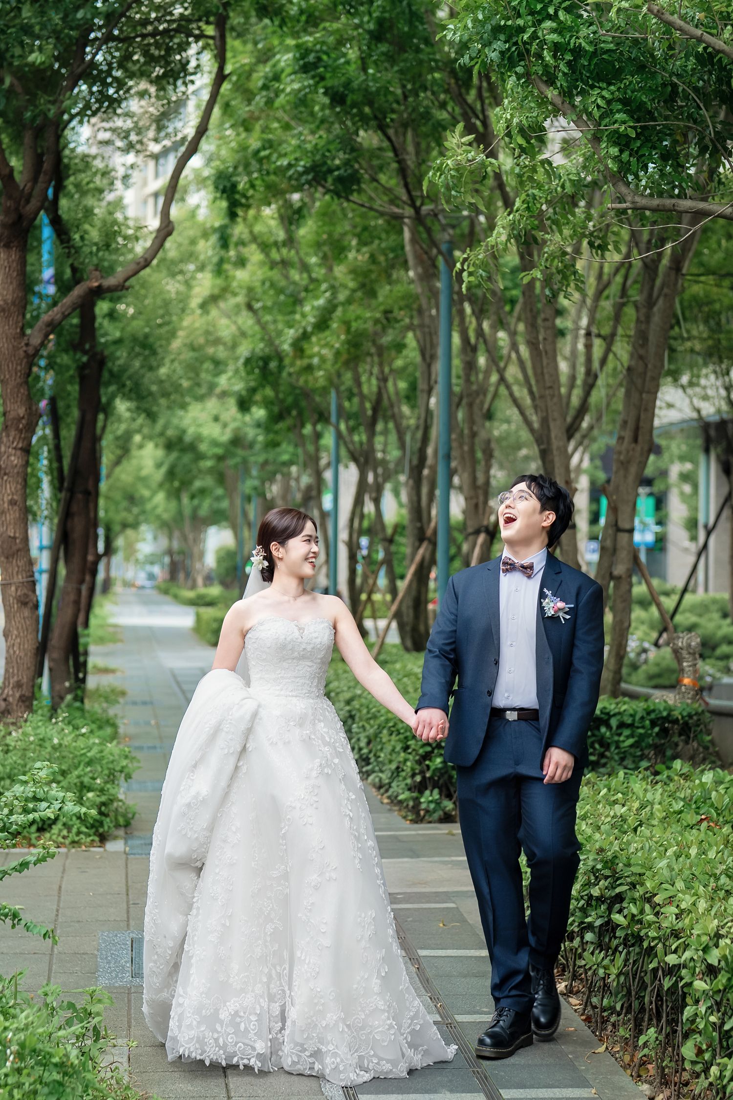 A couple shares joyful laughter while walking together on a tree-lined path during their wedding photo session.