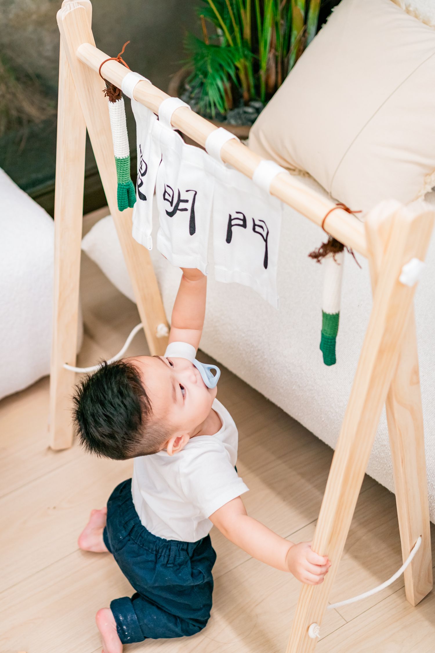 Small child in white shirt and blue shorts reaching up to touch papers hanging on a wooden drying rack indoors.