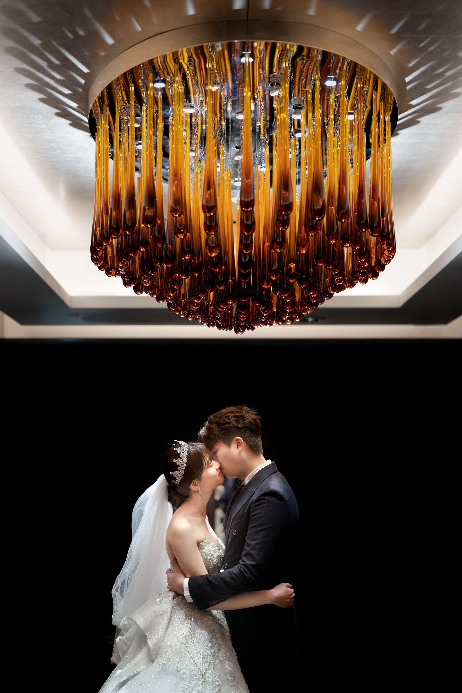 Bride and groom sharing a romantic kiss beneath an elegant amber glass chandelier in a dimly lit wedding venue.