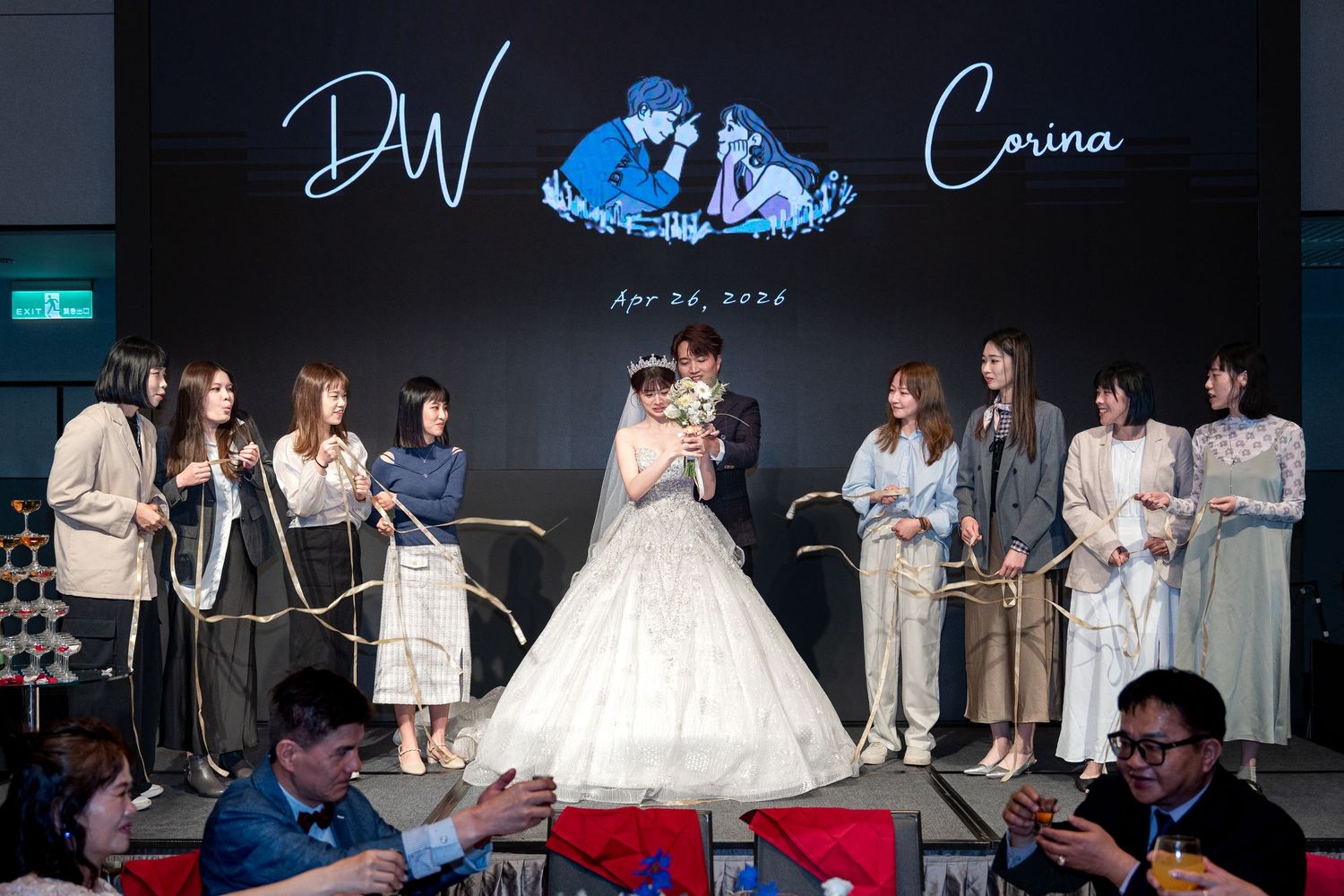 Bride and groom celebrate wedding reception on stage with bridesmaids, backdrop displays names DW and Corina with April 2022 date.