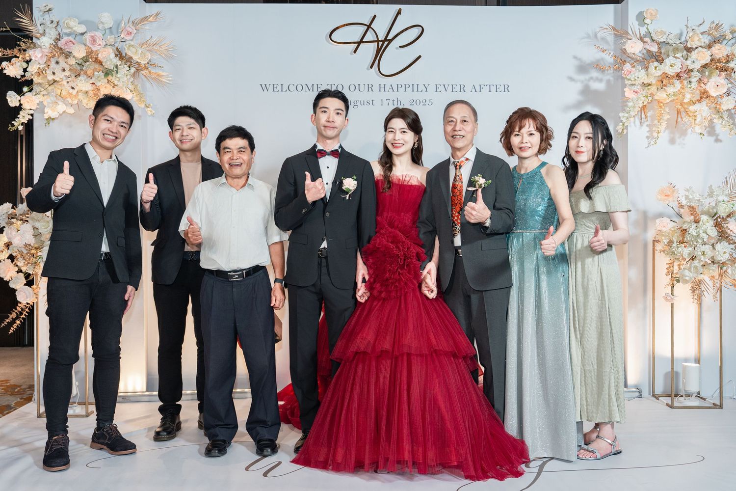 Group of wedding guests in formal attire posing together for photos at an elegant reception venue with light floral decor.