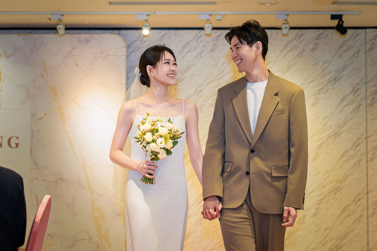 Wedding couple stands together against a marble wall background during their elegant ceremony.