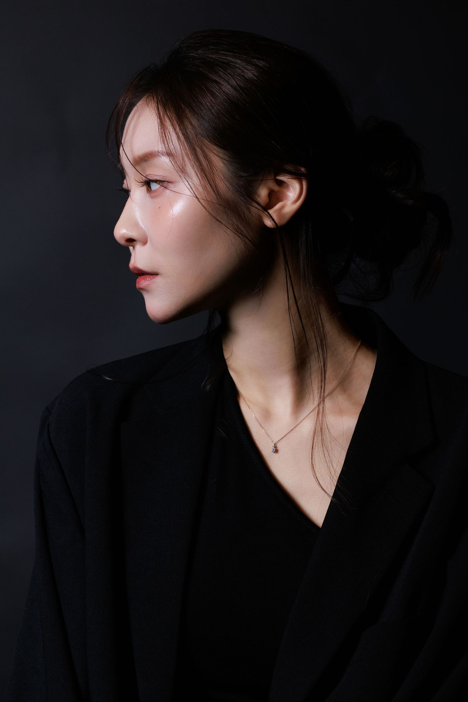 A young Asian woman in a black blazer with a delicate necklace poses in profile against a dark background.