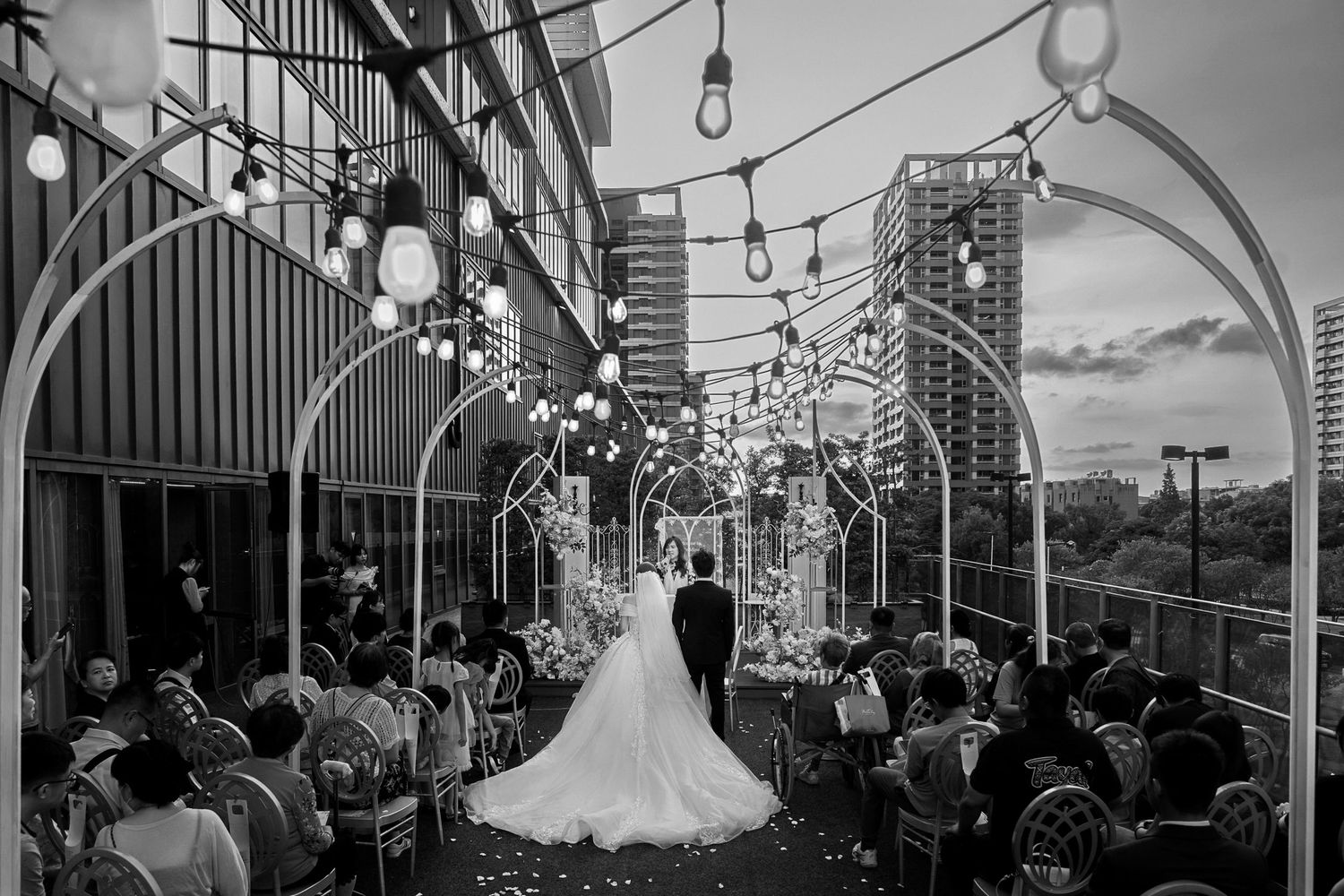 Black and white wedding ceremony on rooftop terrace with string lights, arch decor, bride in gown, groom, and seated guests.