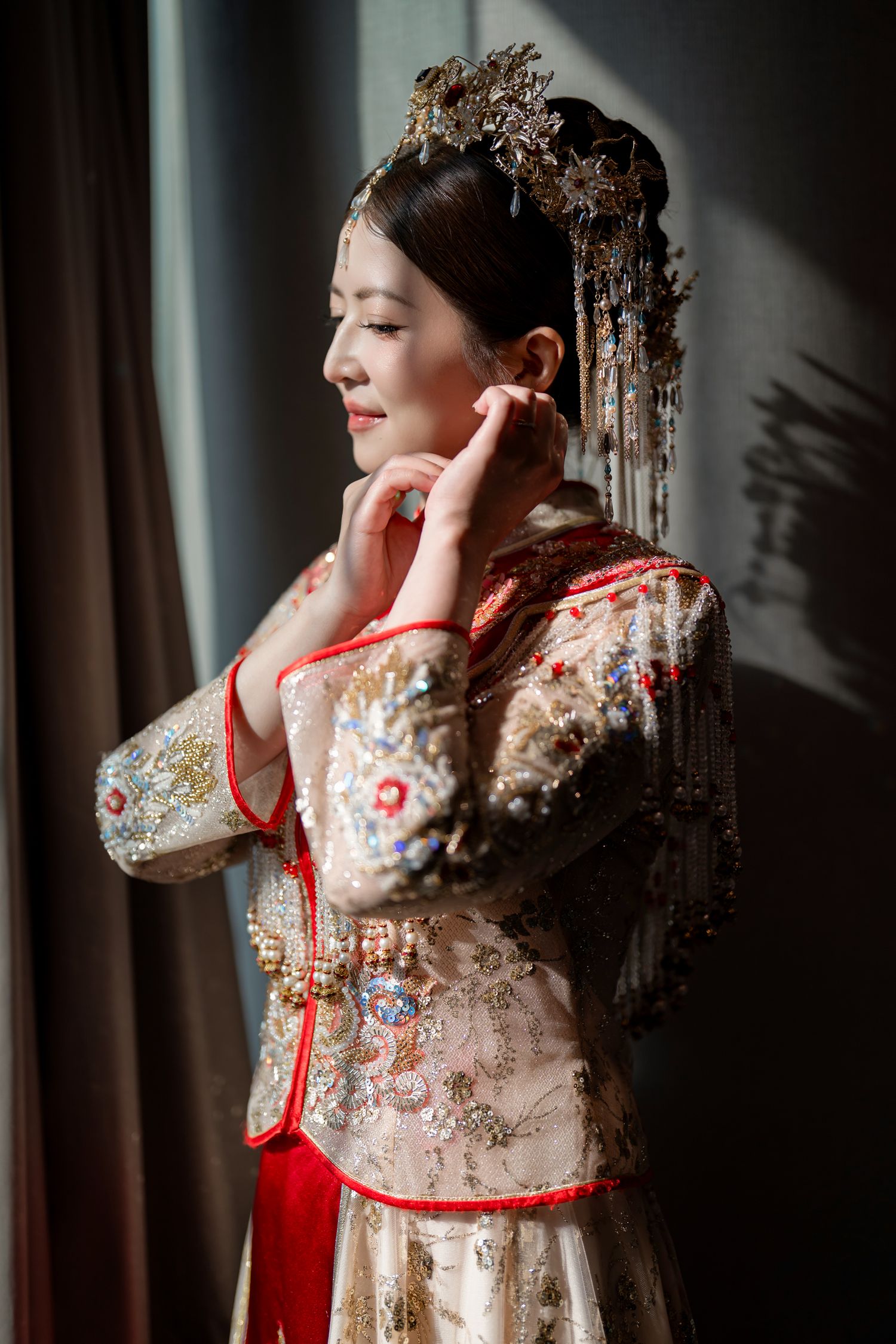 Bride in a red and gold embroidered traditional Chinese wedding gown and ornate gold headpiece adjusts her earring in soft window light.