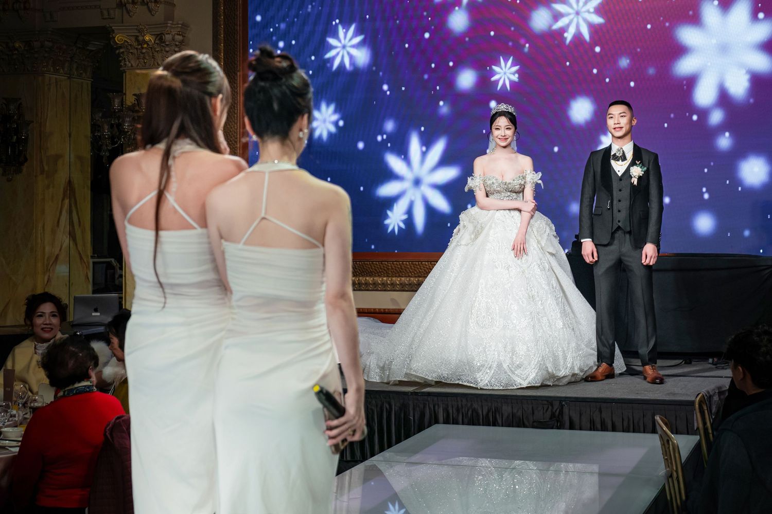 A winter-themed wedding reception features a snowy backdrop with illuminated snowflakes and elegant white decor.