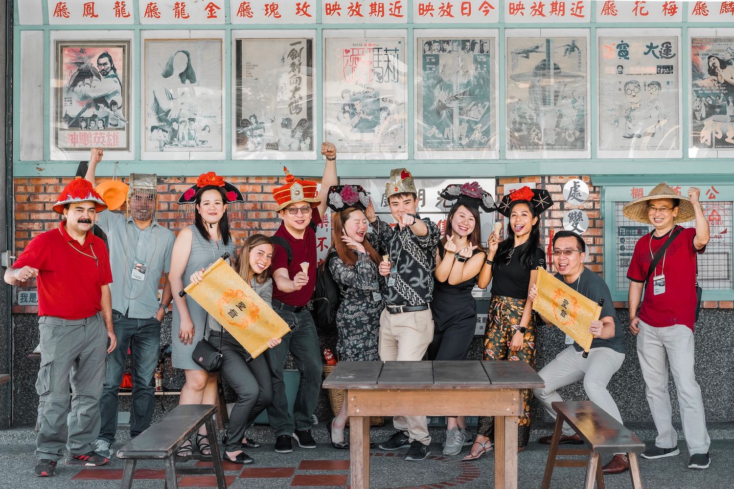 A group stands in front of a wall decorated with traditional Chinese posters, some holding light-colored wooden boxes.