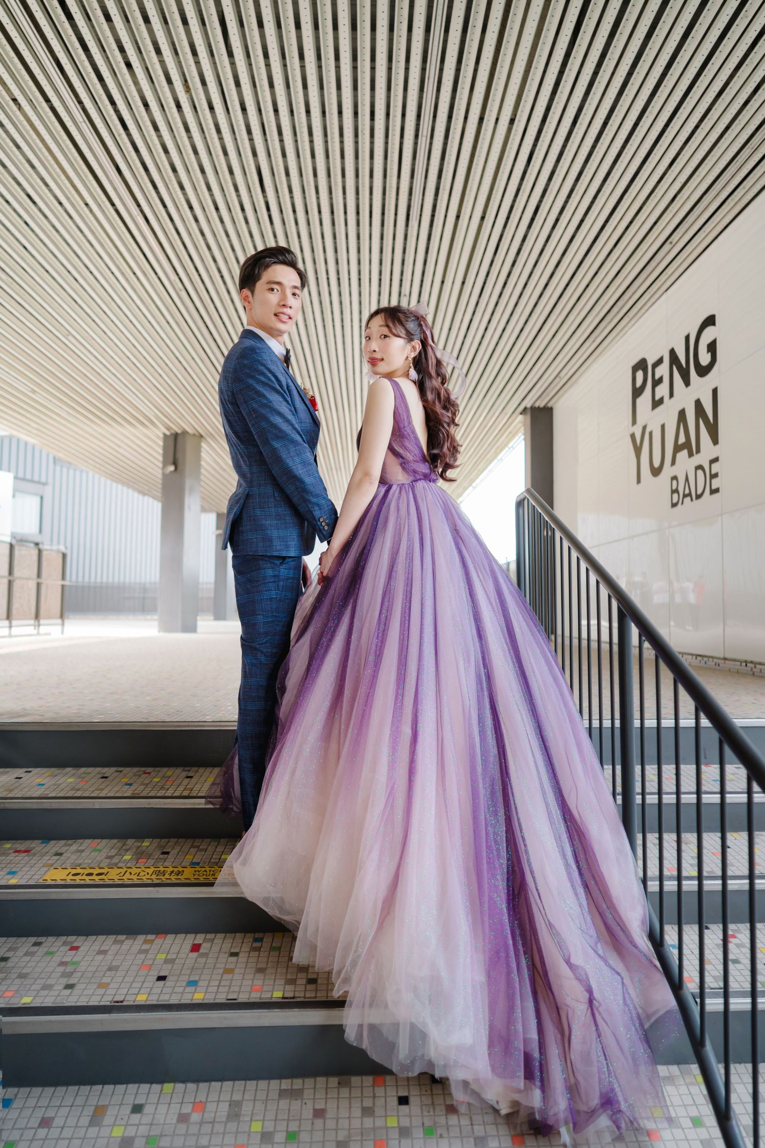 A couple poses on stairs in formal attire, with the woman wearing a flowing purple ombre ball gown and the man in a navy suit.