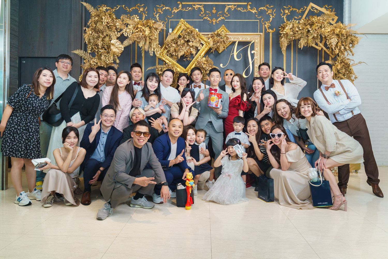A festive group photo of wedding guests posing together beneath an ornate gold decorative arch in an indoor venue.