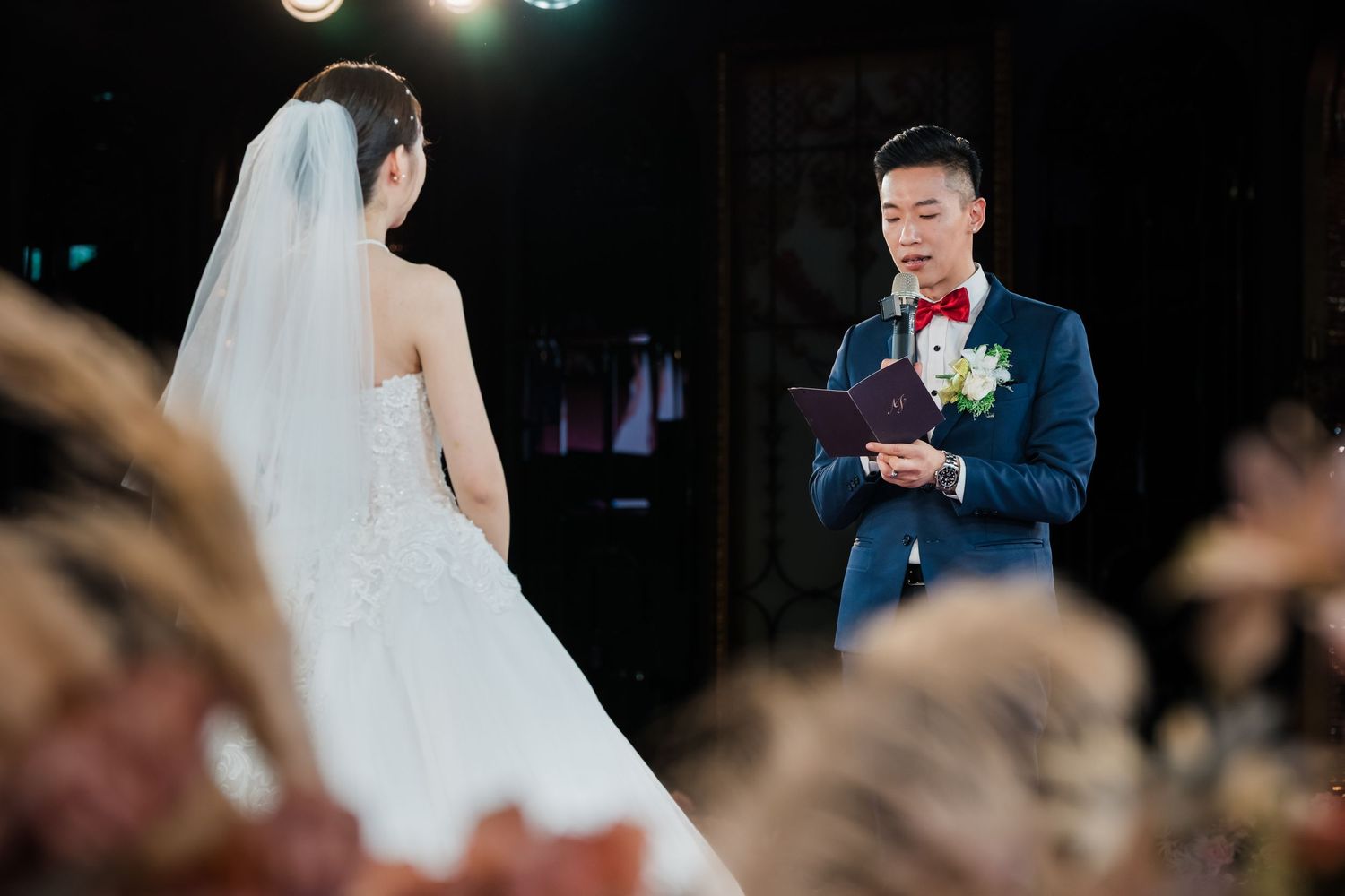A couple exchanges vows during their wedding ceremony with the groom reading from a card while wearing a blue suit.