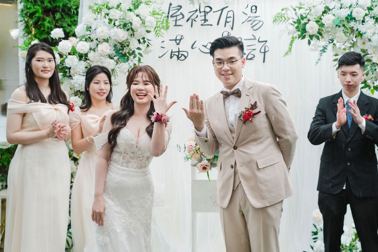 Wedding party celebrates with joyful gestures in front of white floral backdrop at elegant ceremony.