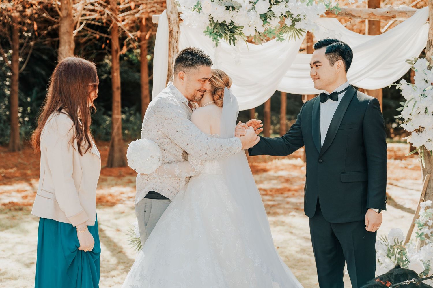 An outdoor wedding ceremony takes place under a white fabric arch with fall foliage in the background.