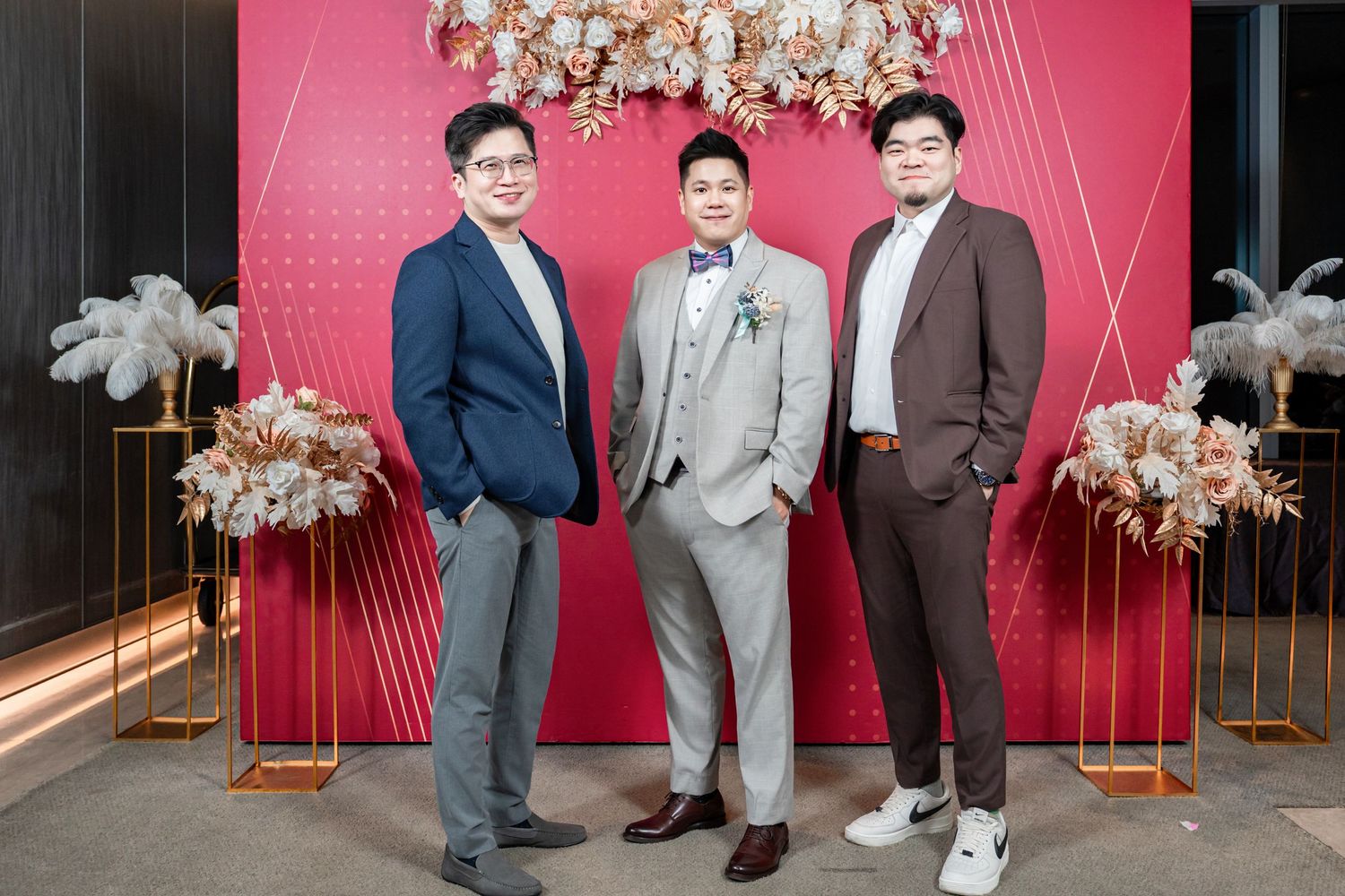 Three people in formal attire stand together against a pink backdrop with floral decorations at an event venue.