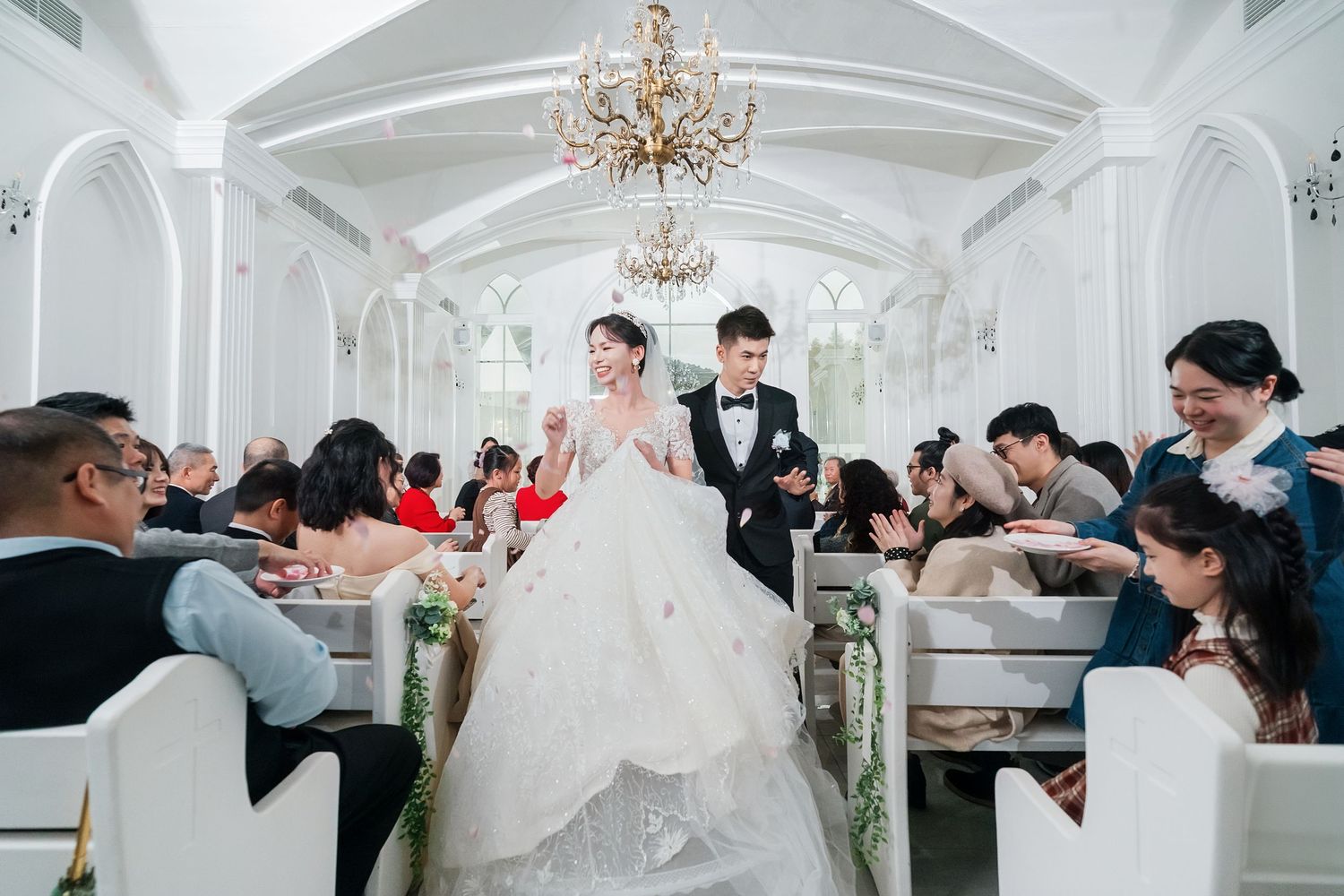 Panoramic view of wedding guests reacting emotionally in an elegant white ballroom with crystal chandeliers.