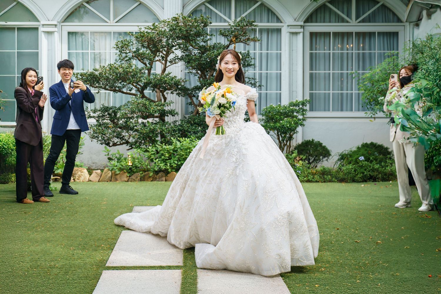 Wedding party members toss flower petals at a couple during an outdoor garden celebration.