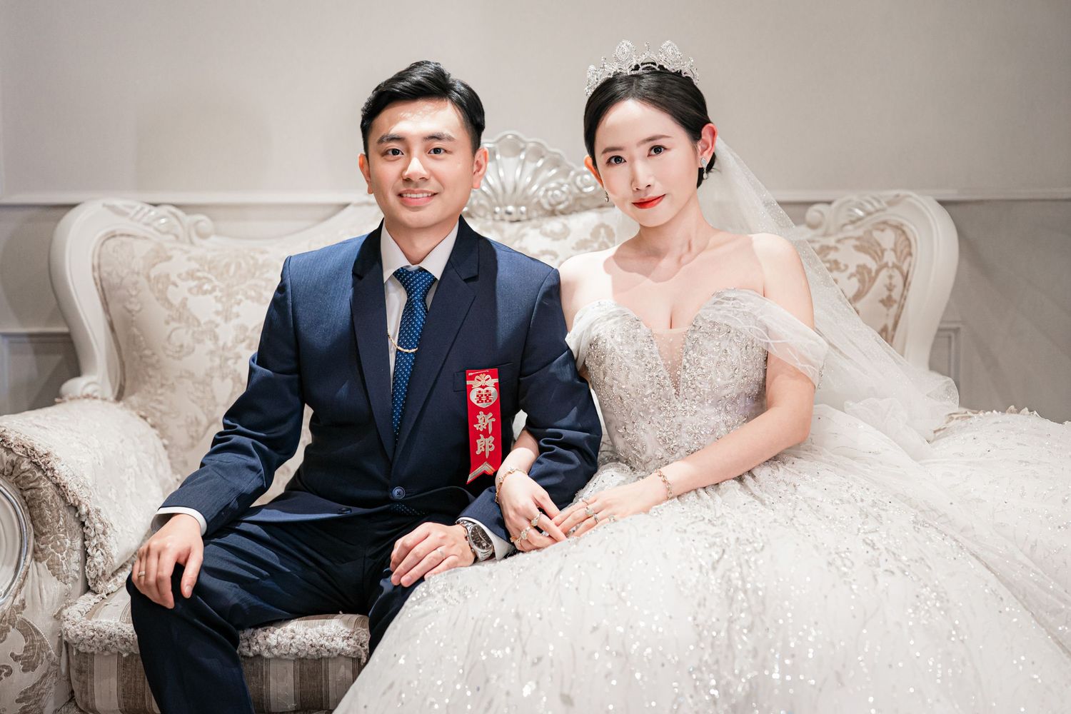 A bride in a white wedding dress and groom in navy suit pose together on an elegant beige sofa.