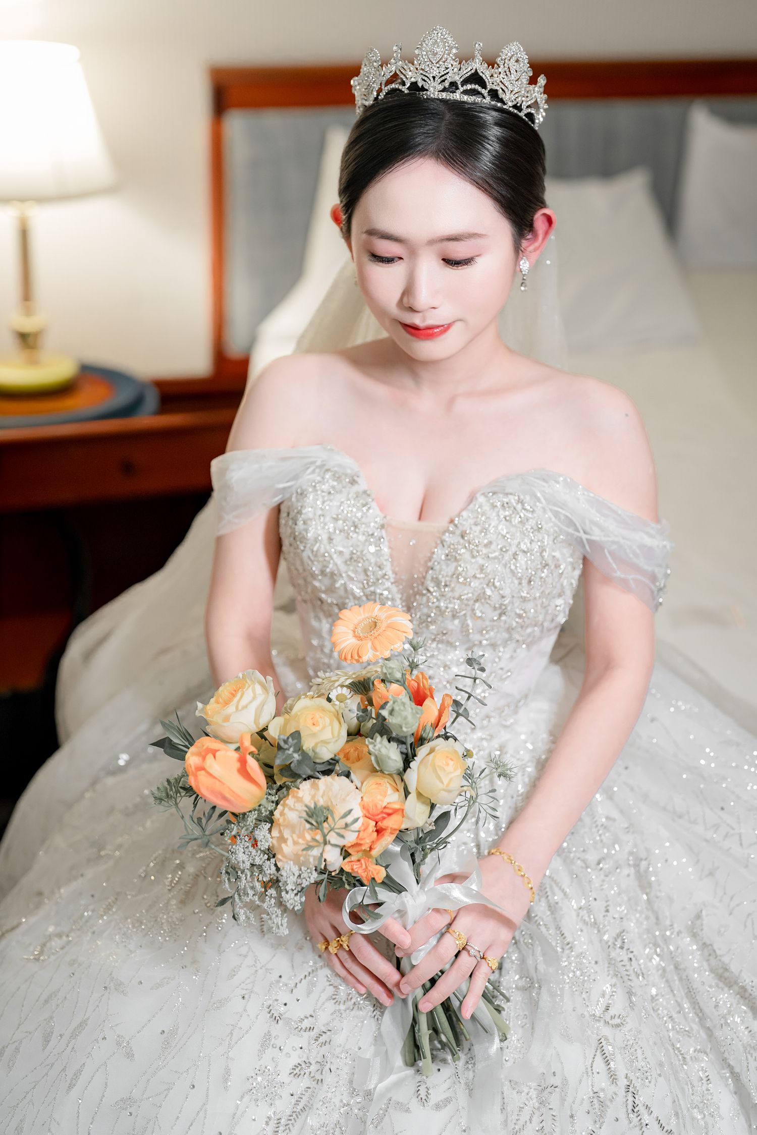 Elegant bride in off-shoulder lace gown with crystal crown holds peach rose bouquet in soft indoor lighting.
