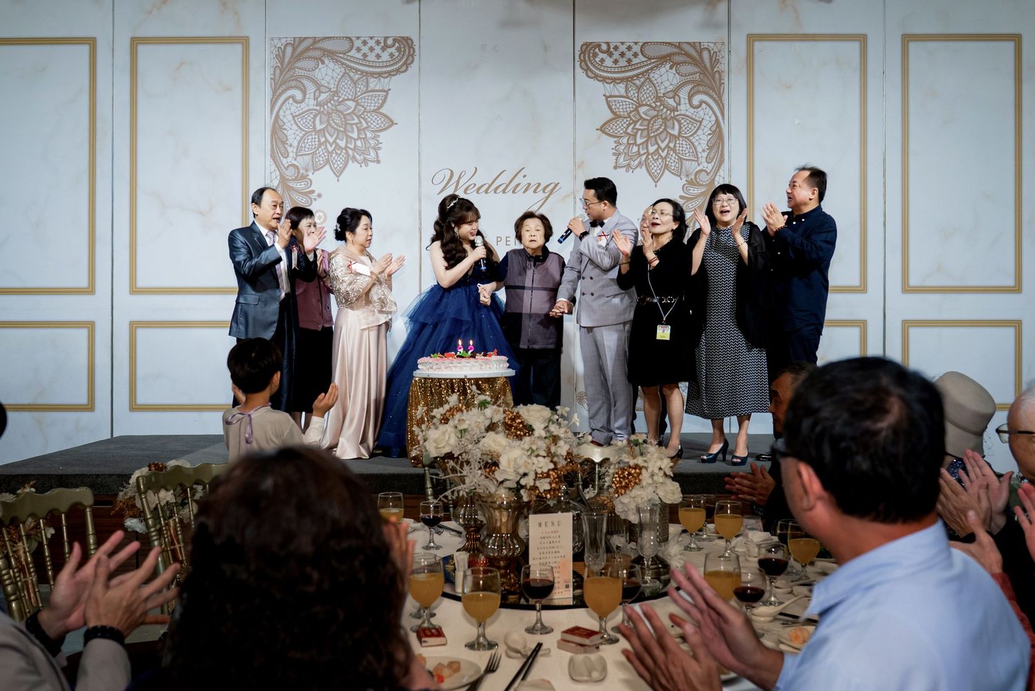 Wedding guests stand and applaud during an elegant reception with ornate wall panels and decorated dining tables.