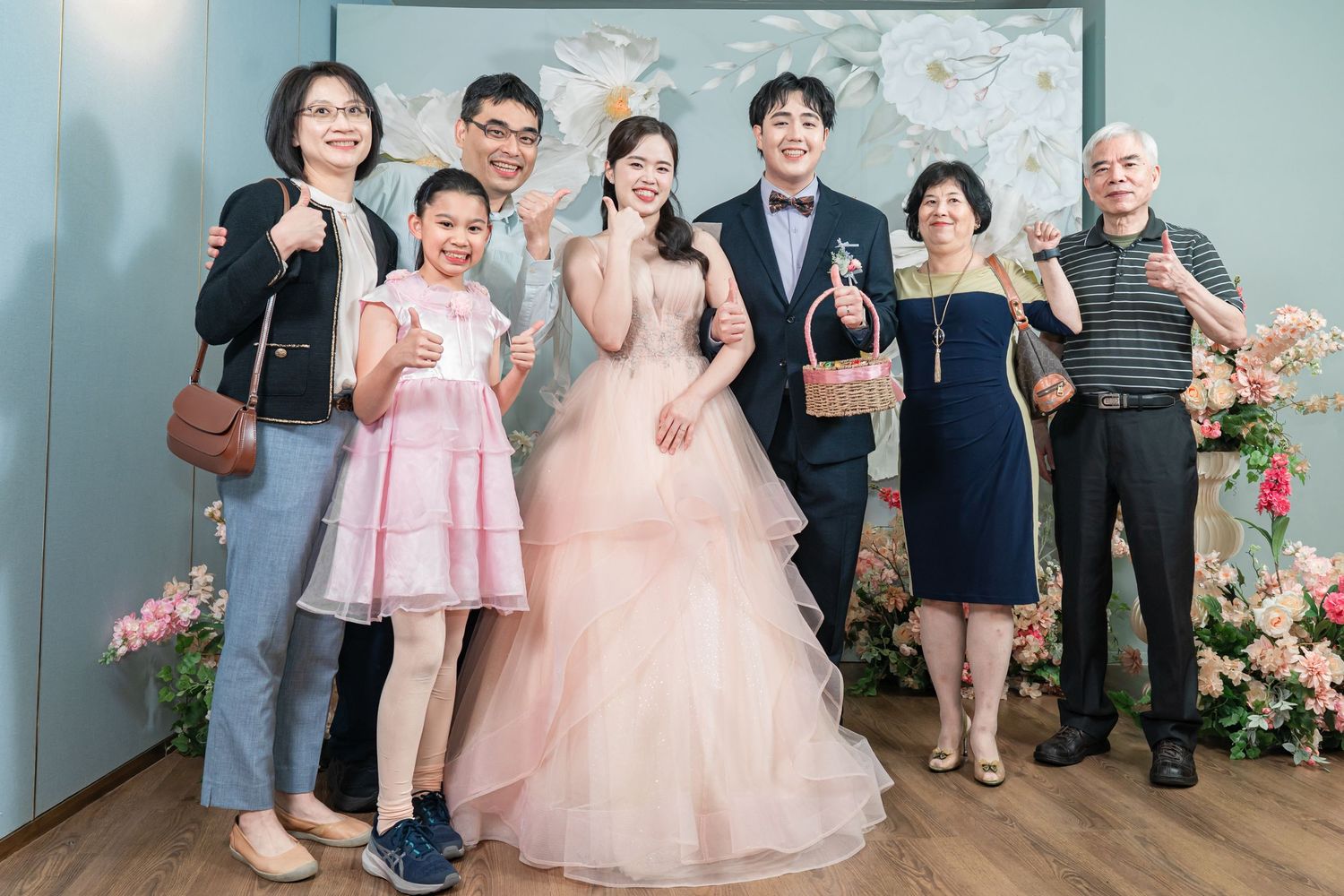 A group poses together at a wedding celebration with the bride wearing a pink princess gown among floral decorations.