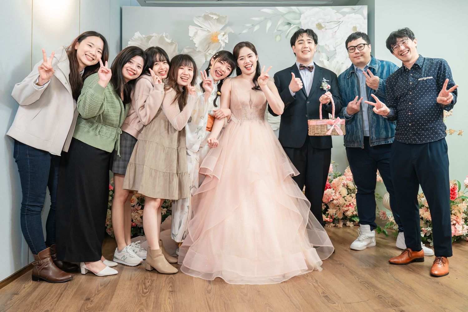Group photo of wedding attendees sitting together on floor with pink floral decorations in background.