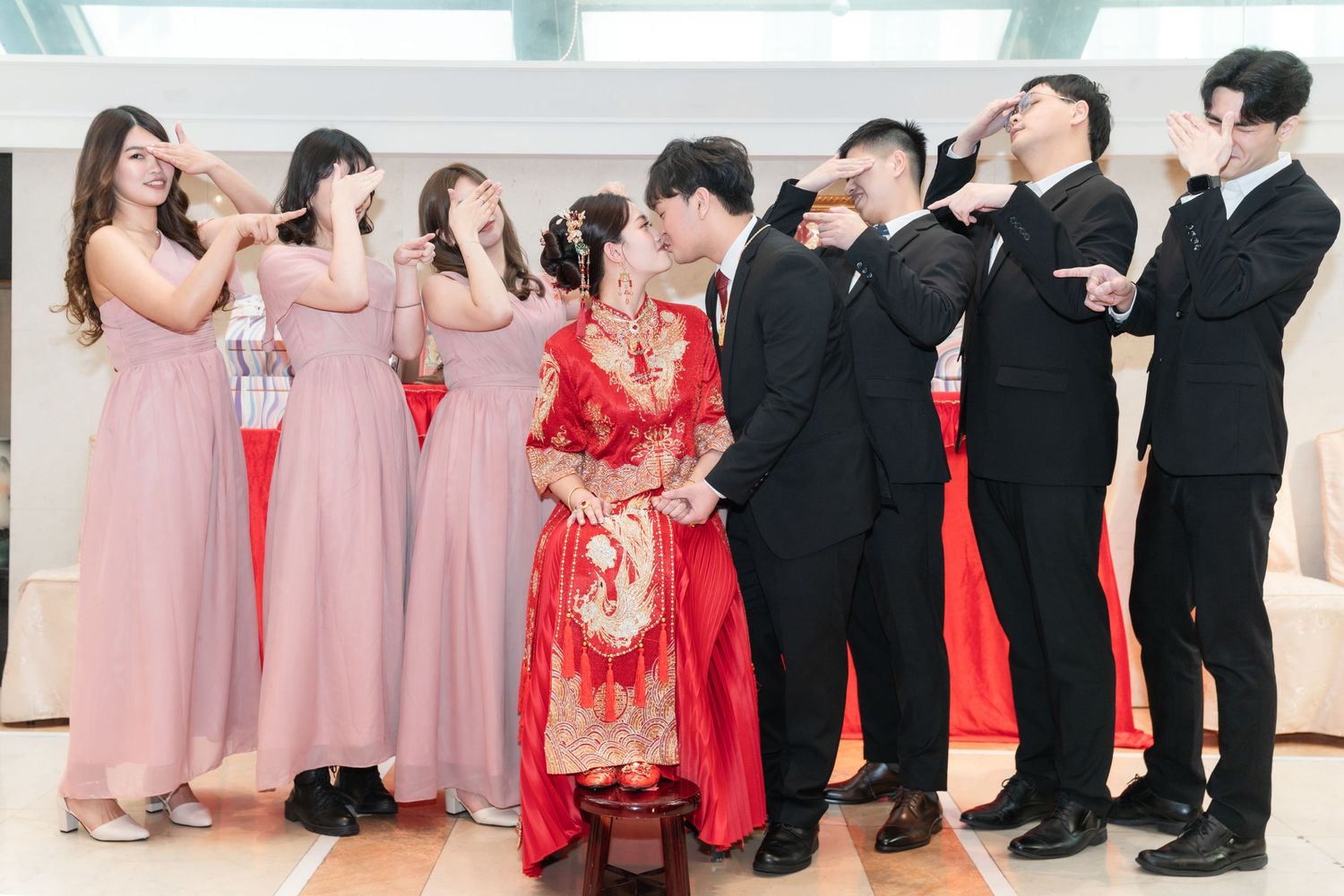 Wedding party lined up with bridesmaids in pink dresses and bride in traditional red qipao dress standing with groomsmen in black suits.